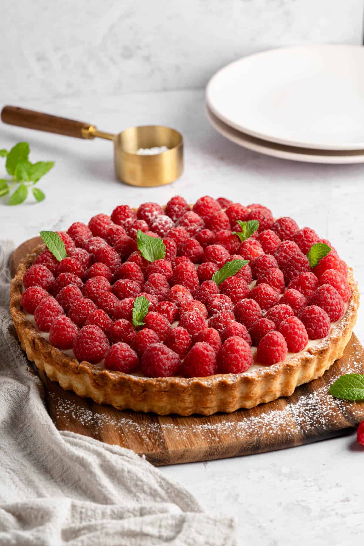 A fresh raspberry tart with plates and a measuring cup behind it.