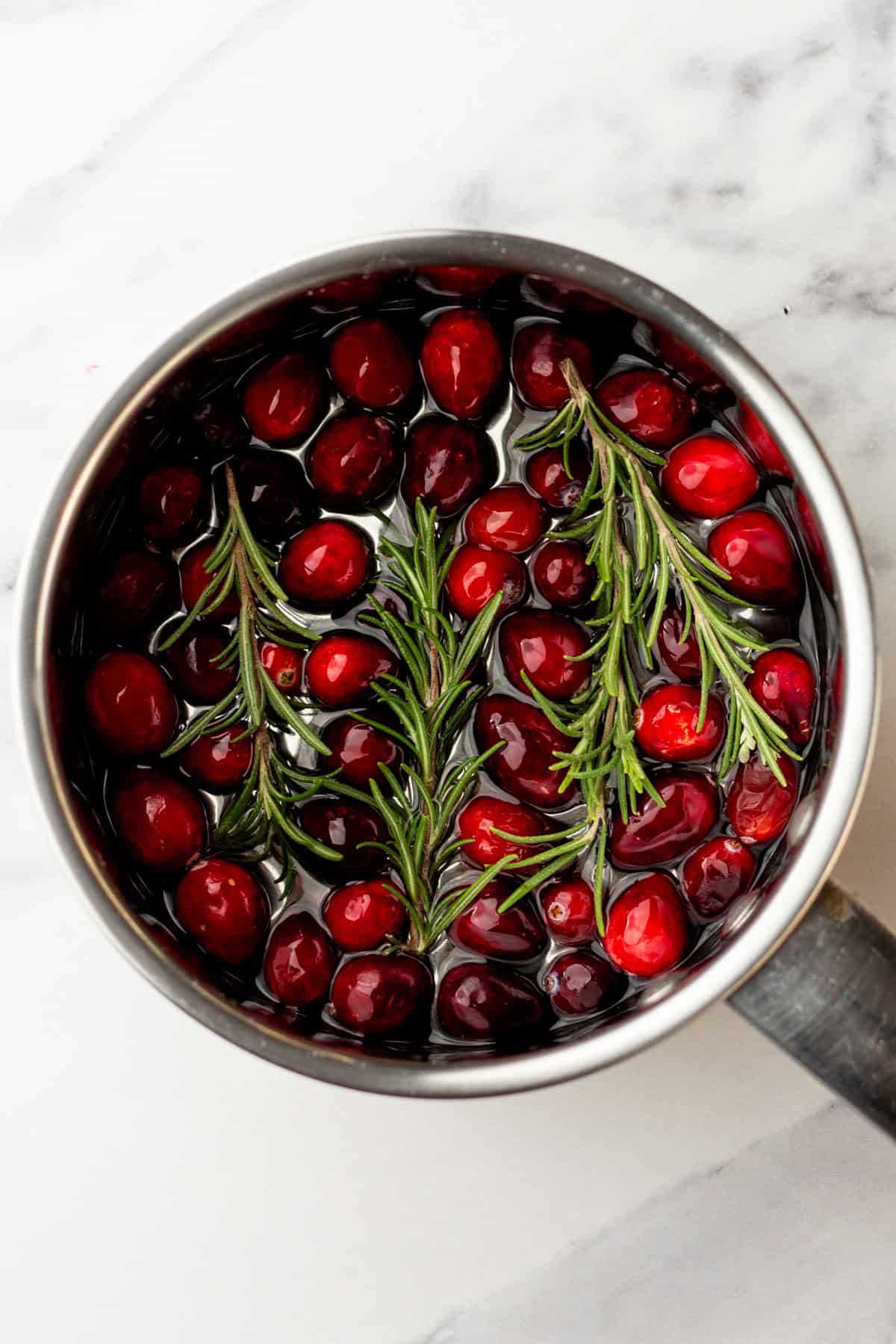 Combining fresh cranberries and rosemary sprigs in a pot with sugar and water.