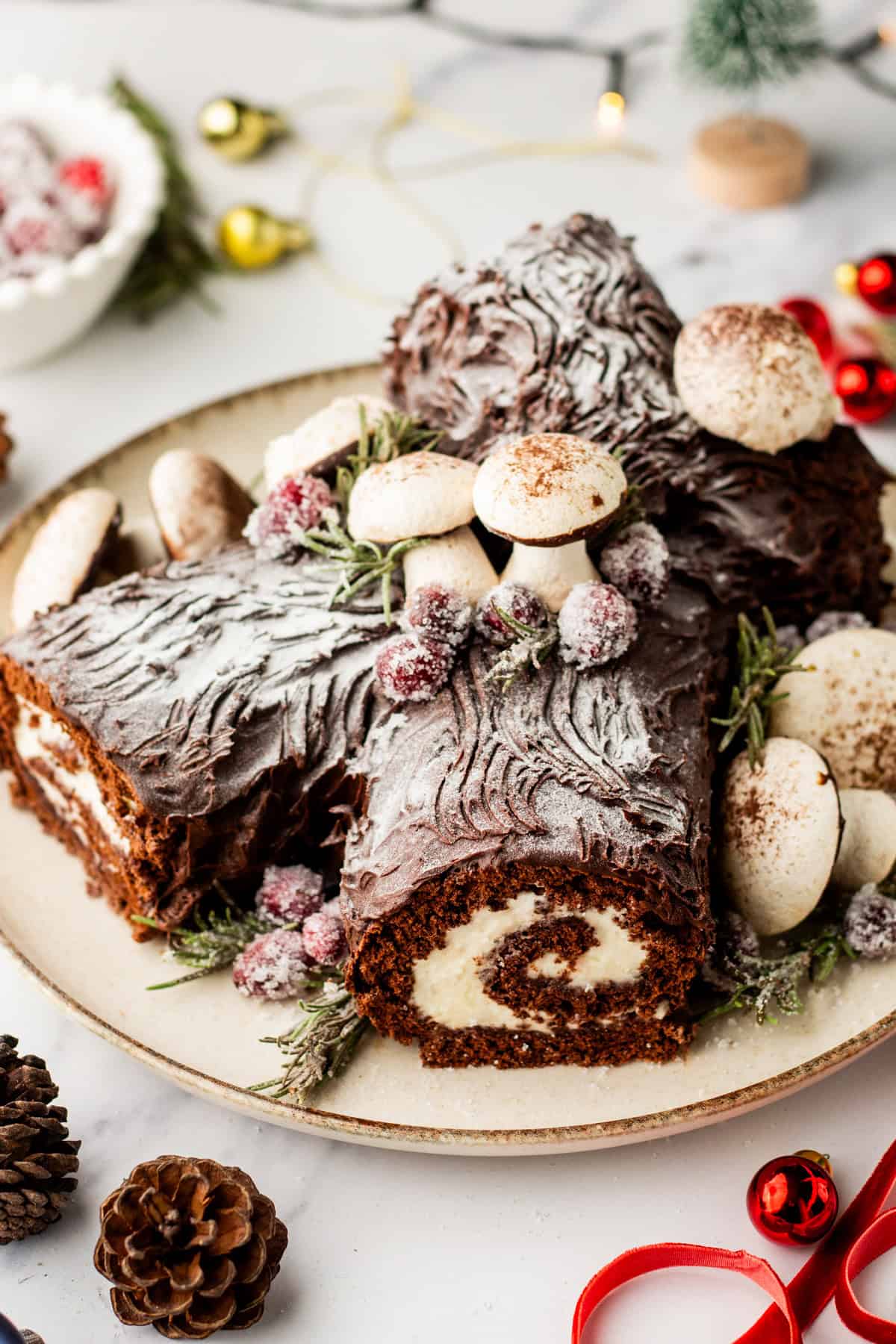 A decorated buche de noel on a plate with meringue mushrooms and candied cranberries and rosemary.