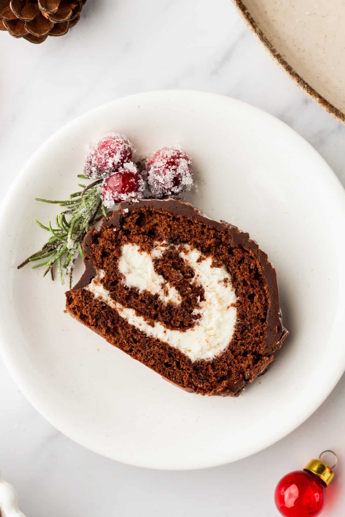 A slice of buche de noel on a white plate with sugared cranberries and sugared rosemary.