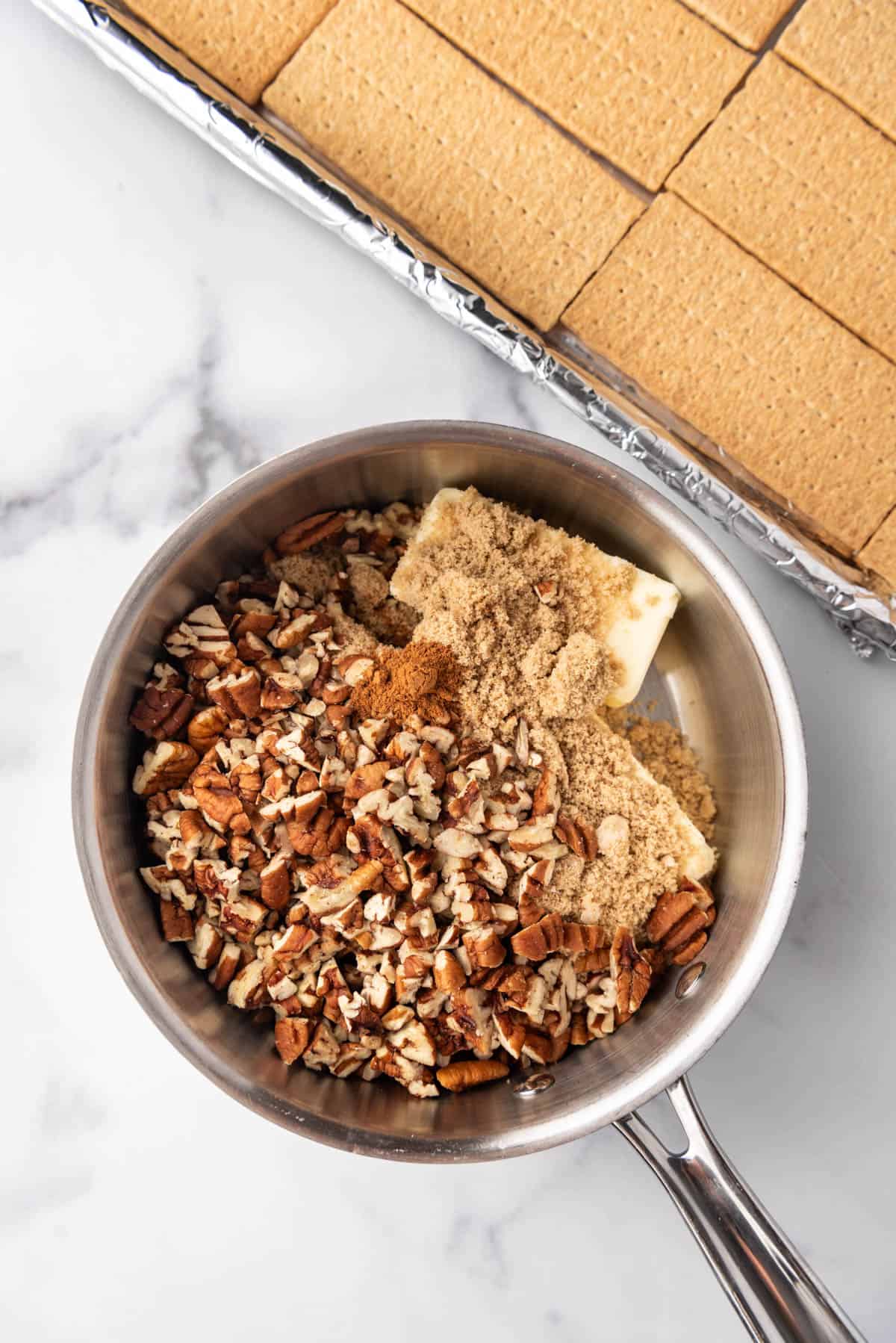 Combining butter, brown sugar, and chopped pecans in a medium saucepan next to a baking sheet of graham crackers.