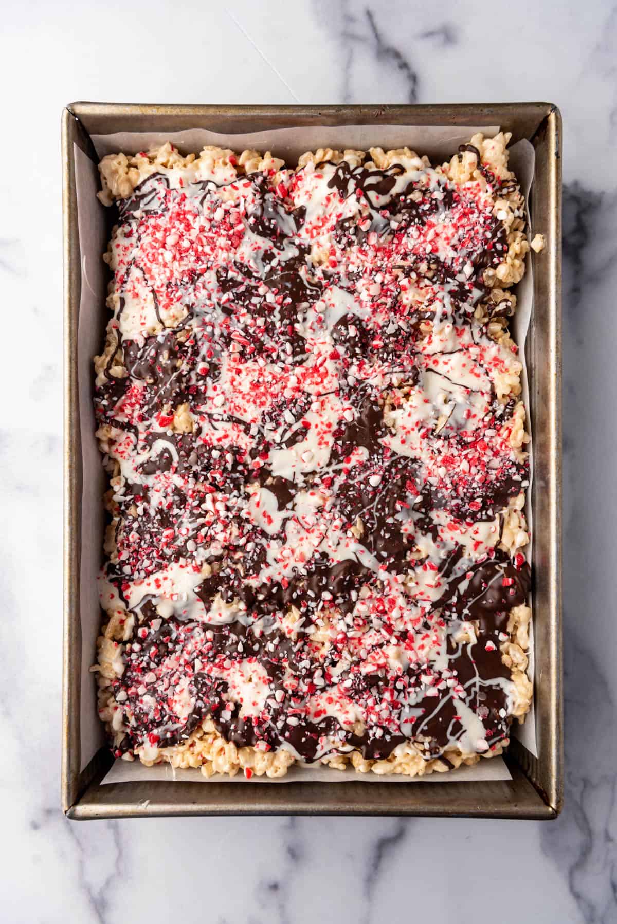 Melted white and dark chocolate drizzled over rice krispies treats with crushed candy cane pieces sprinkled on top.