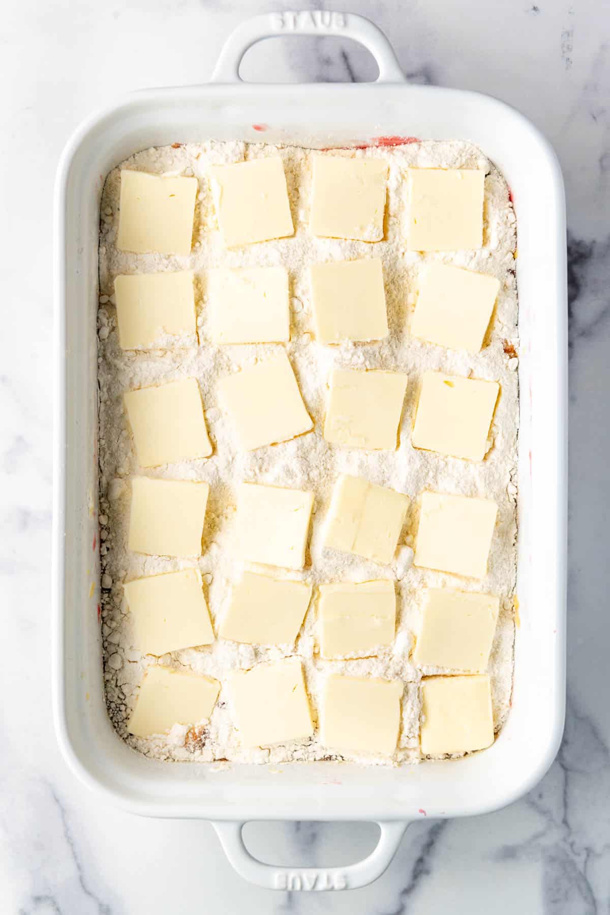 Sliced butter arranged on top of cake mix and fruit in a white baking dish.