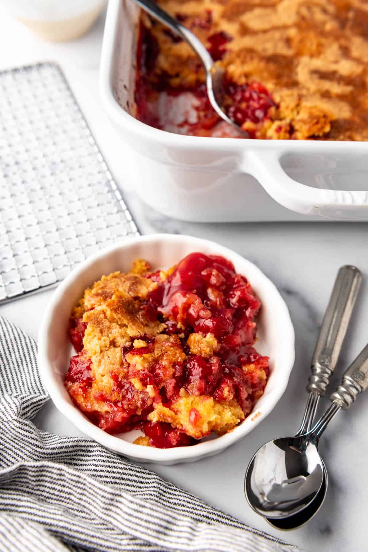 A white bowl of cherry pineapple dump cake next to a white baking dish.