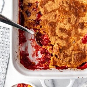 An overhead image of a serving spoon scooping out cherry pineapple dump cake from a white baking dish.