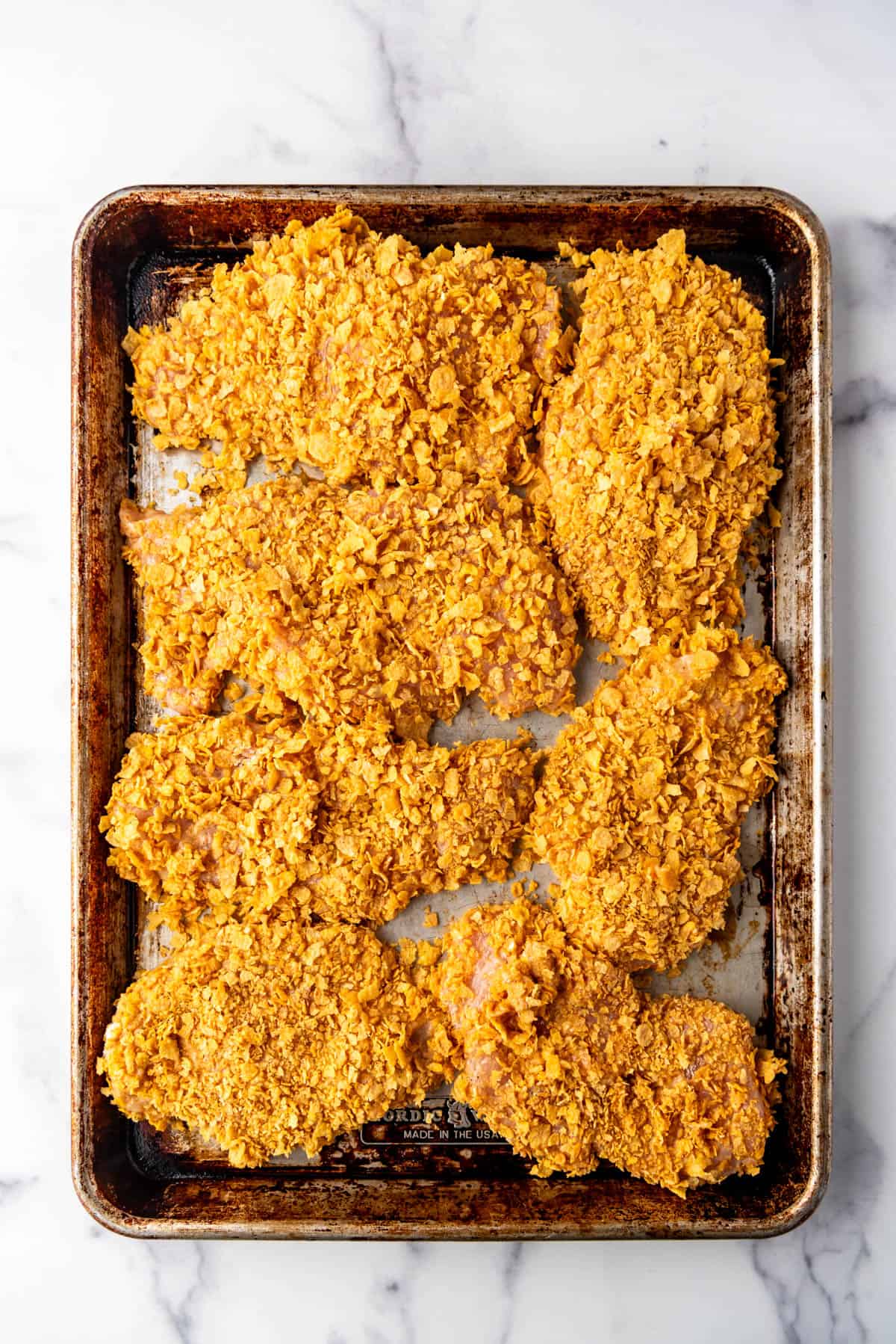 Chicken breasts breaded with cornflakes on a baking sheet.