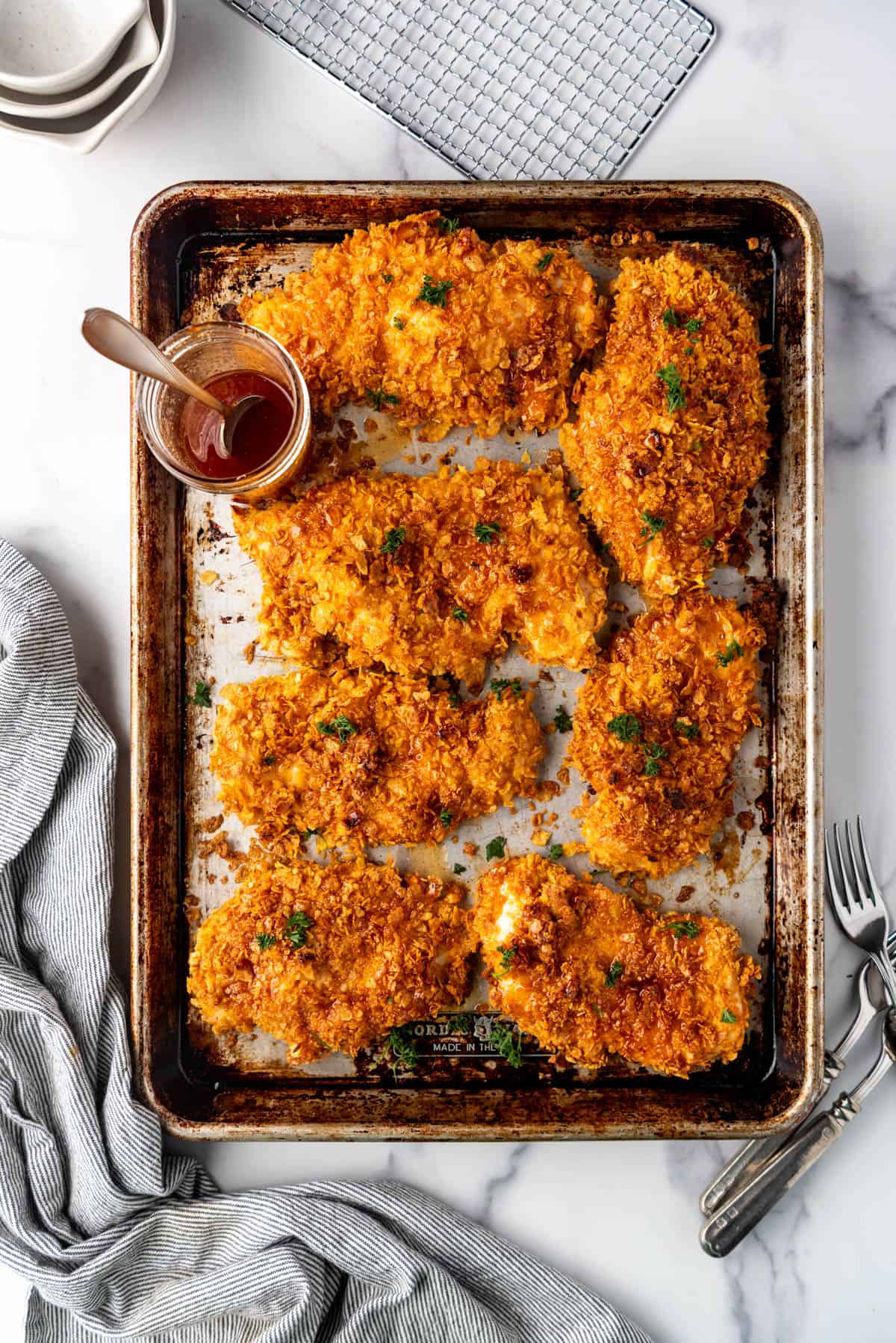 An overhead image of a baking sheet filled with cornflake chicken and a jar of homemade hot honey sauce.