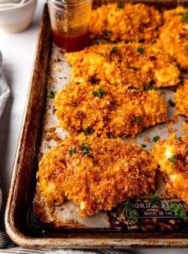 An image of crispy oven baked cornflake chicken with hot honey sauce on a baking sheet.