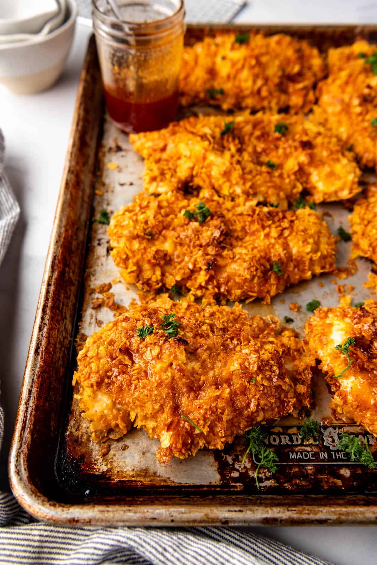 An image of crispy oven baked cornflake chicken with hot honey sauce on a baking sheet.