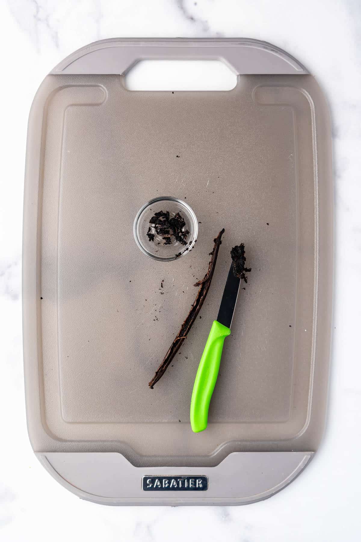 A paring knife and a vanilla bean pod on a cutting board with the seeds scraped out of the vanilla bean pod.