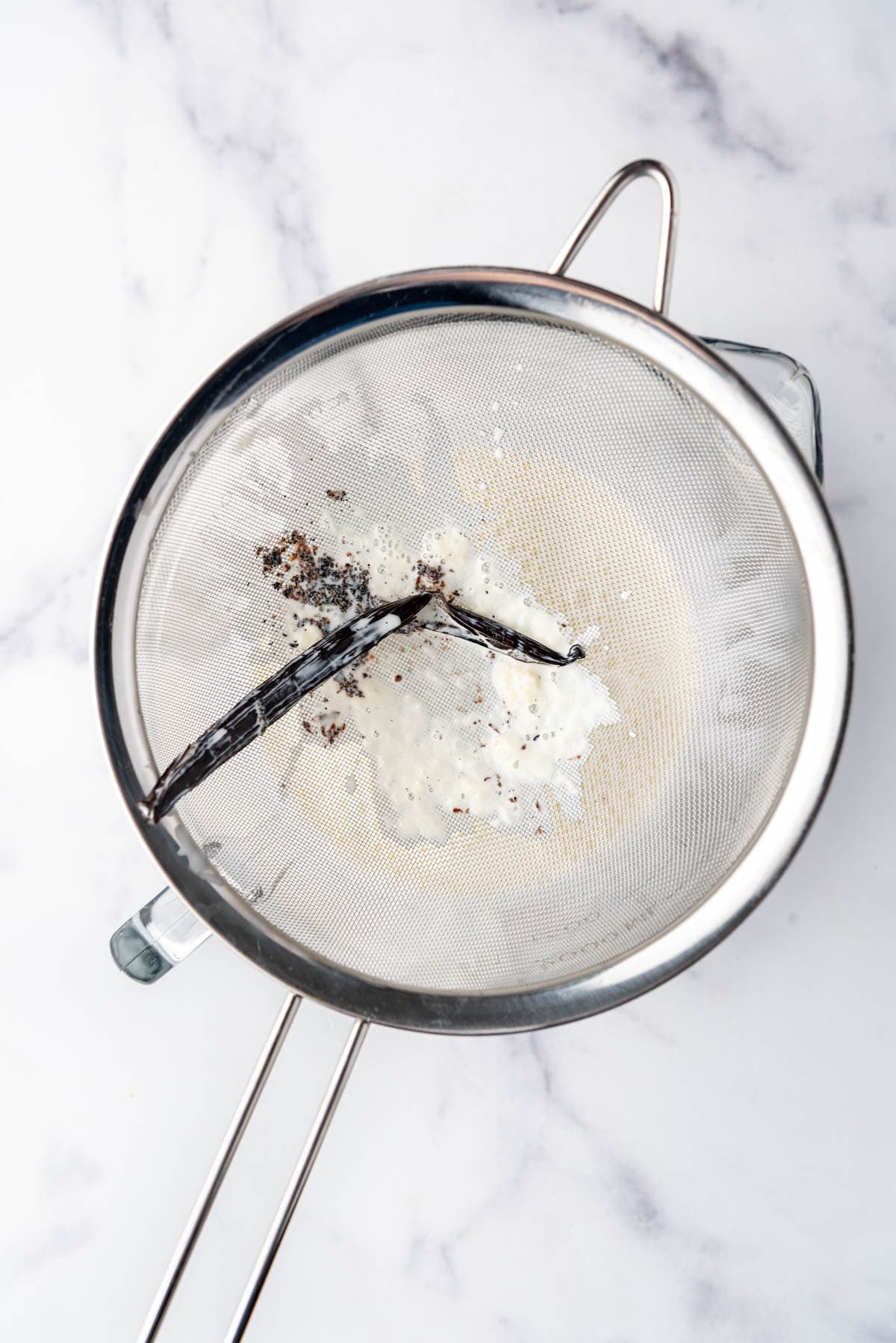 A vanilla bean pod and the remnants of cooked cream, sugar, and egg in a fine mesh sieve over a glass bowl.