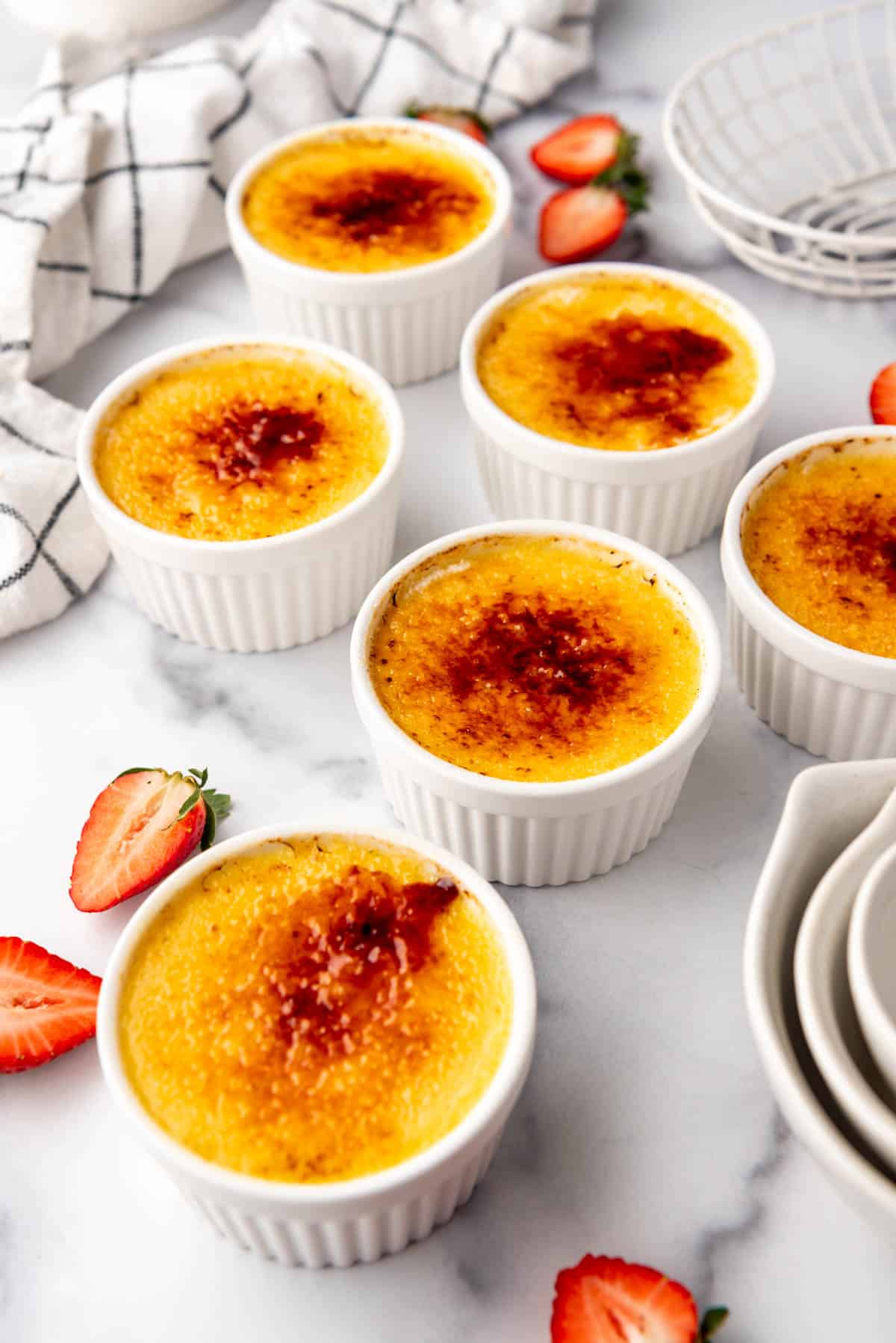 Images of homemade creme brulee in white ramekins with sliced strawberry halves scattered nearby.