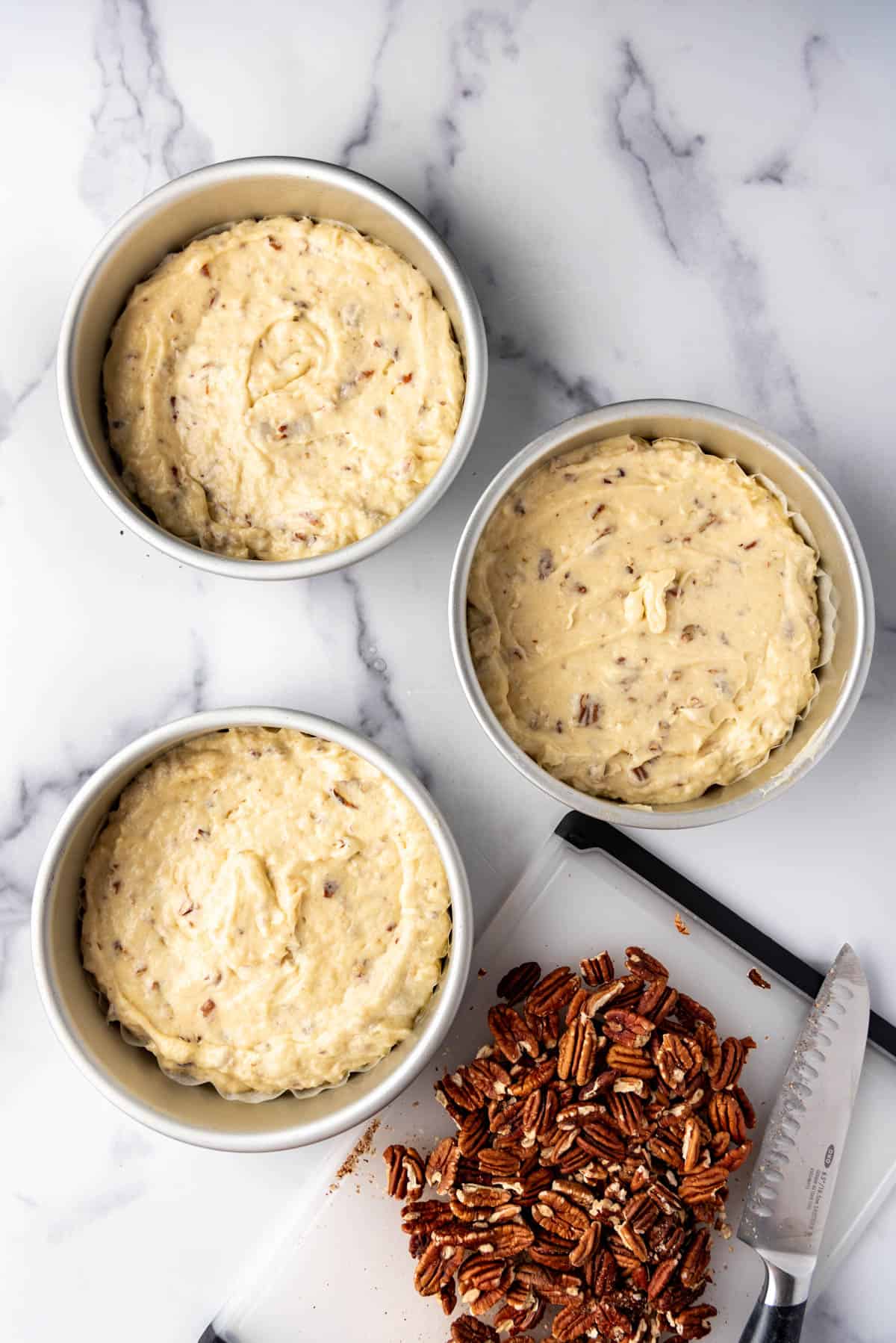 An overhead image of three cake pans filled with Italian cream cake batter next to chopped pecans on a   cutting board with a large knife.