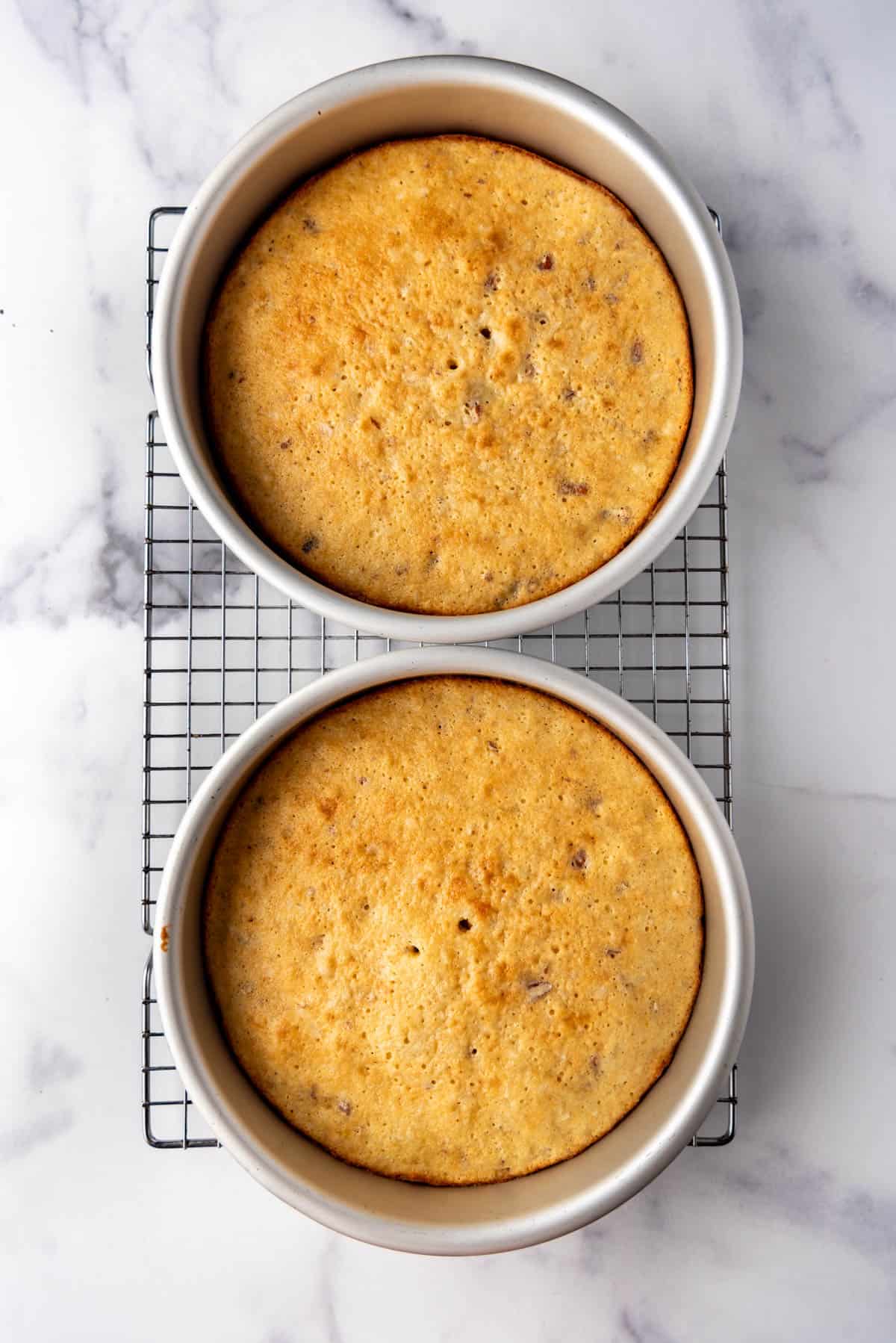 Two baked cake layers in pans on a wire cooling rack.