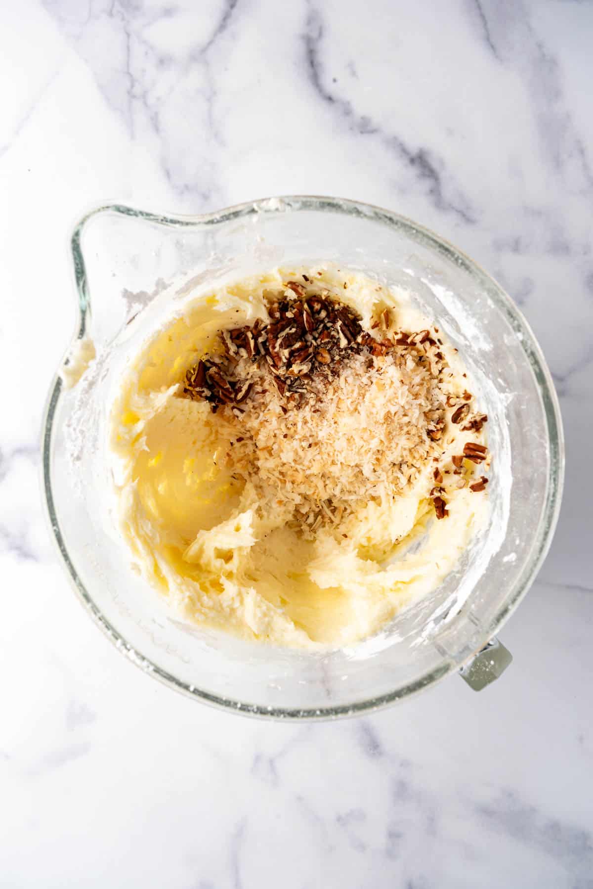 Adding toasted coconut and chopped toasted pecans to cream cheese frosting in a glass mixing bowl.