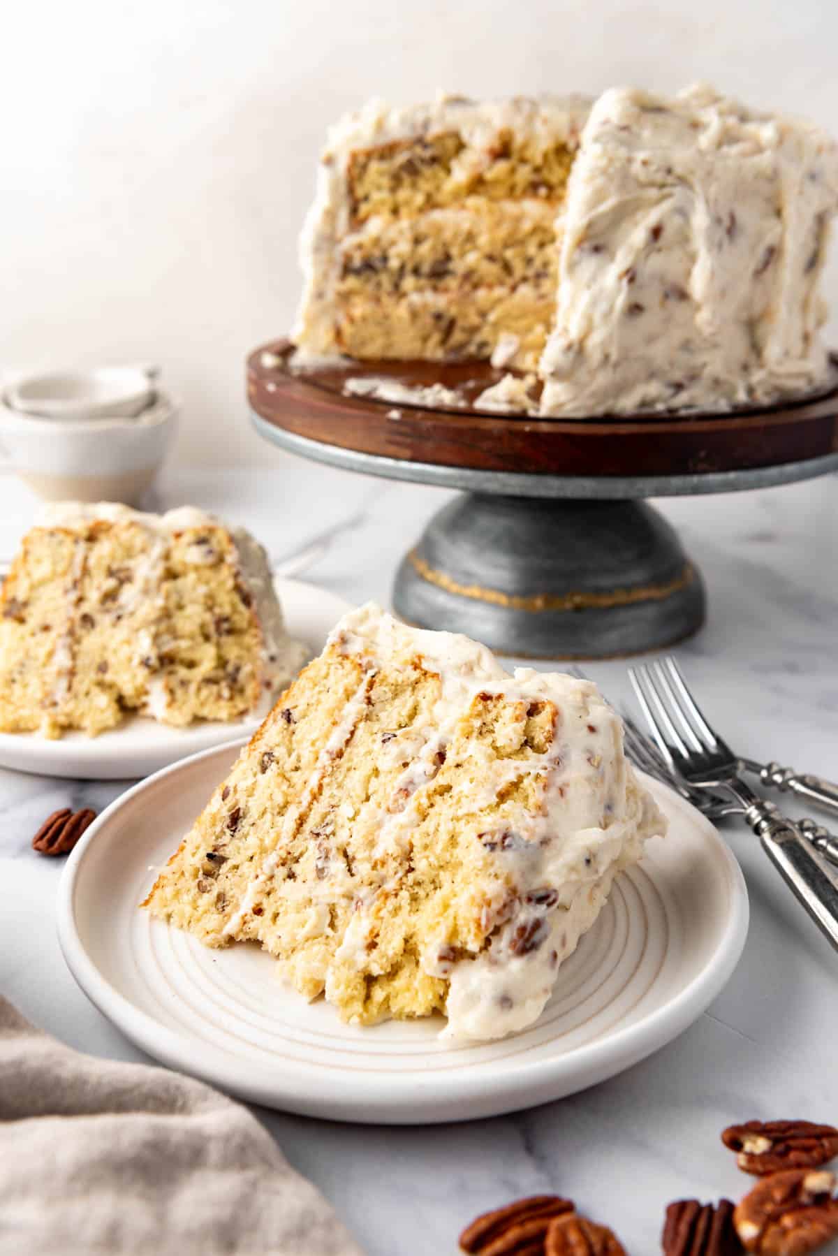 Une tranche de gâteau crème italienne sur une assiette devant le reste du gâteau sur un support à gâteau.