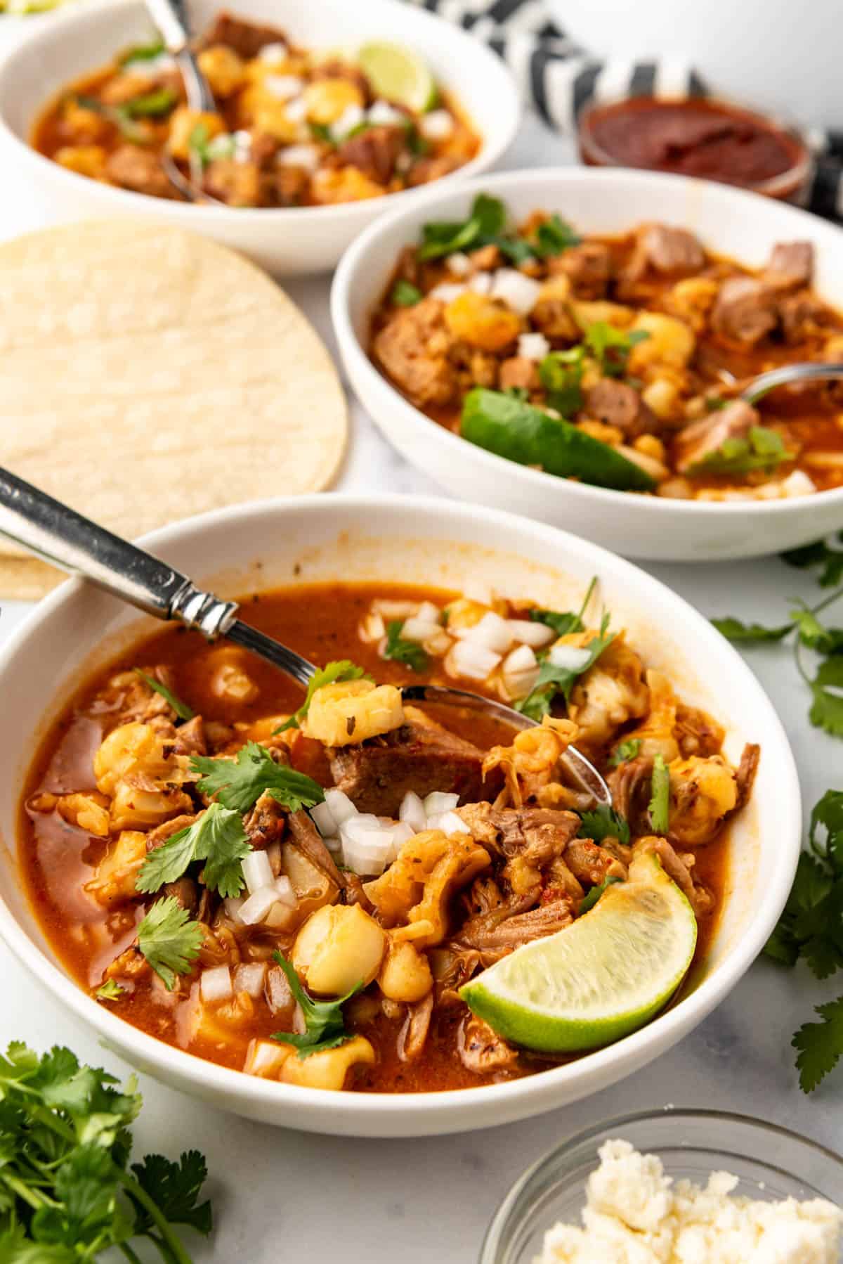 Bowls of red posole with garnishes of fresh cilantro, chopped white onion, and lime wedges on top.