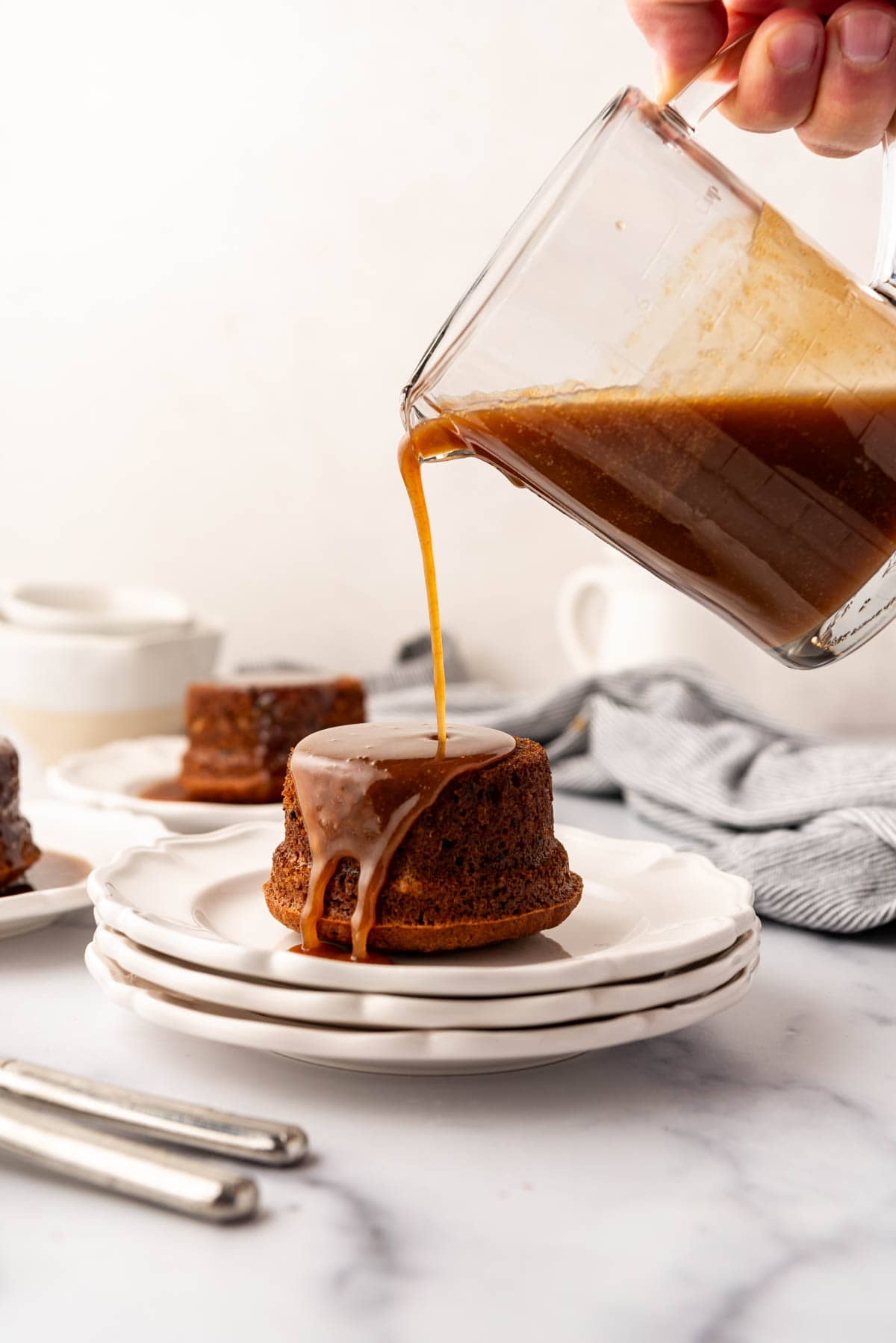 An image of warm toffee sauce being poured over an individual serving of sticky toffee pudding on a white plate stacked on top of two other dessert plates.