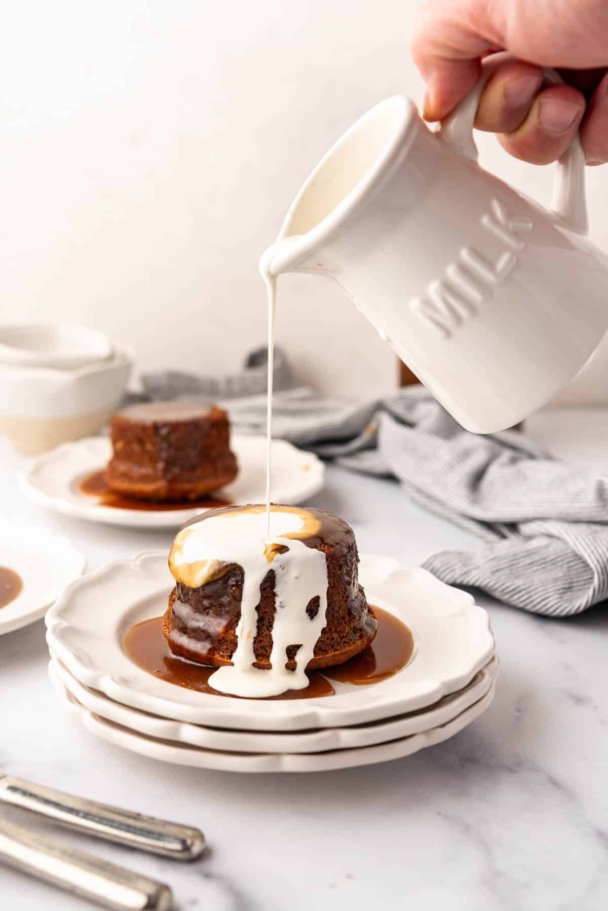 An image of cream being poured over a serving of sticky toffee pudding on dessert plates.