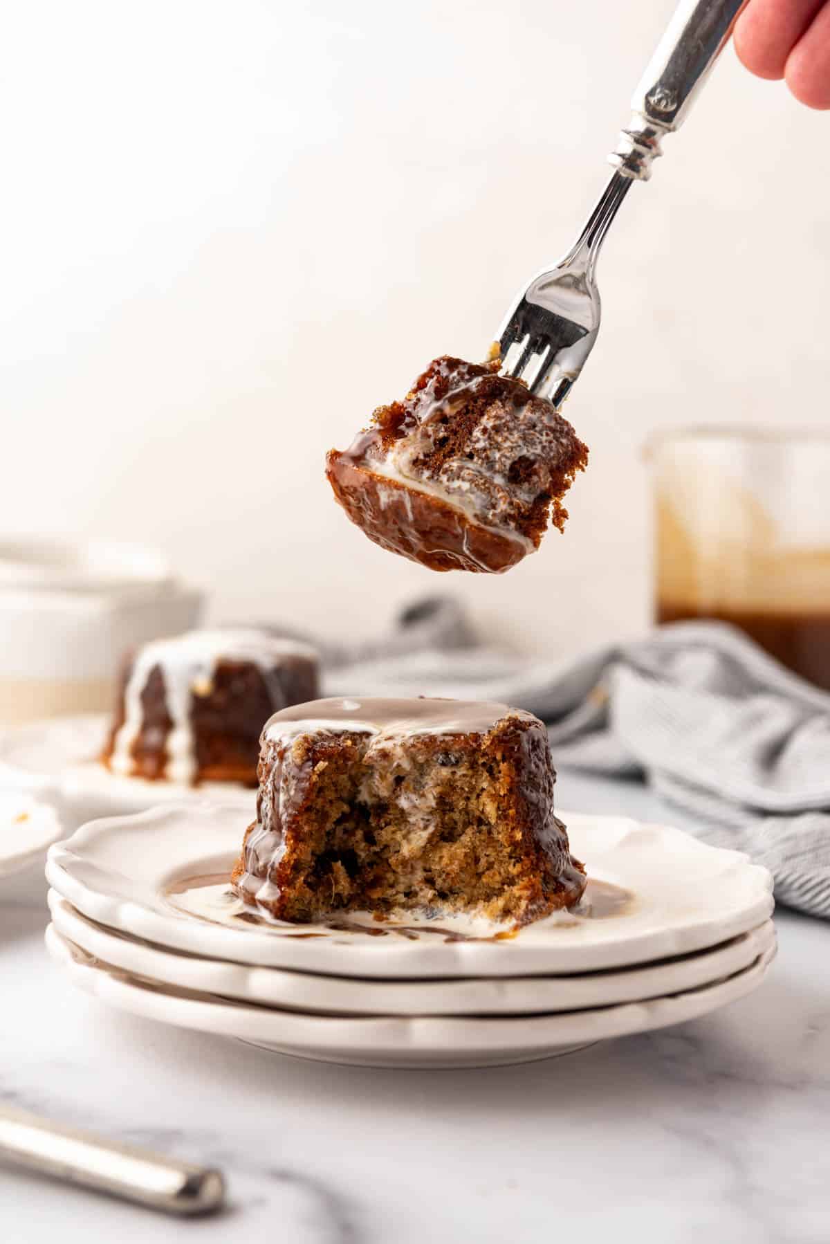 An image of a fork holding a bite of sticky toffee pudding over the rest of the cake on dessert plates.