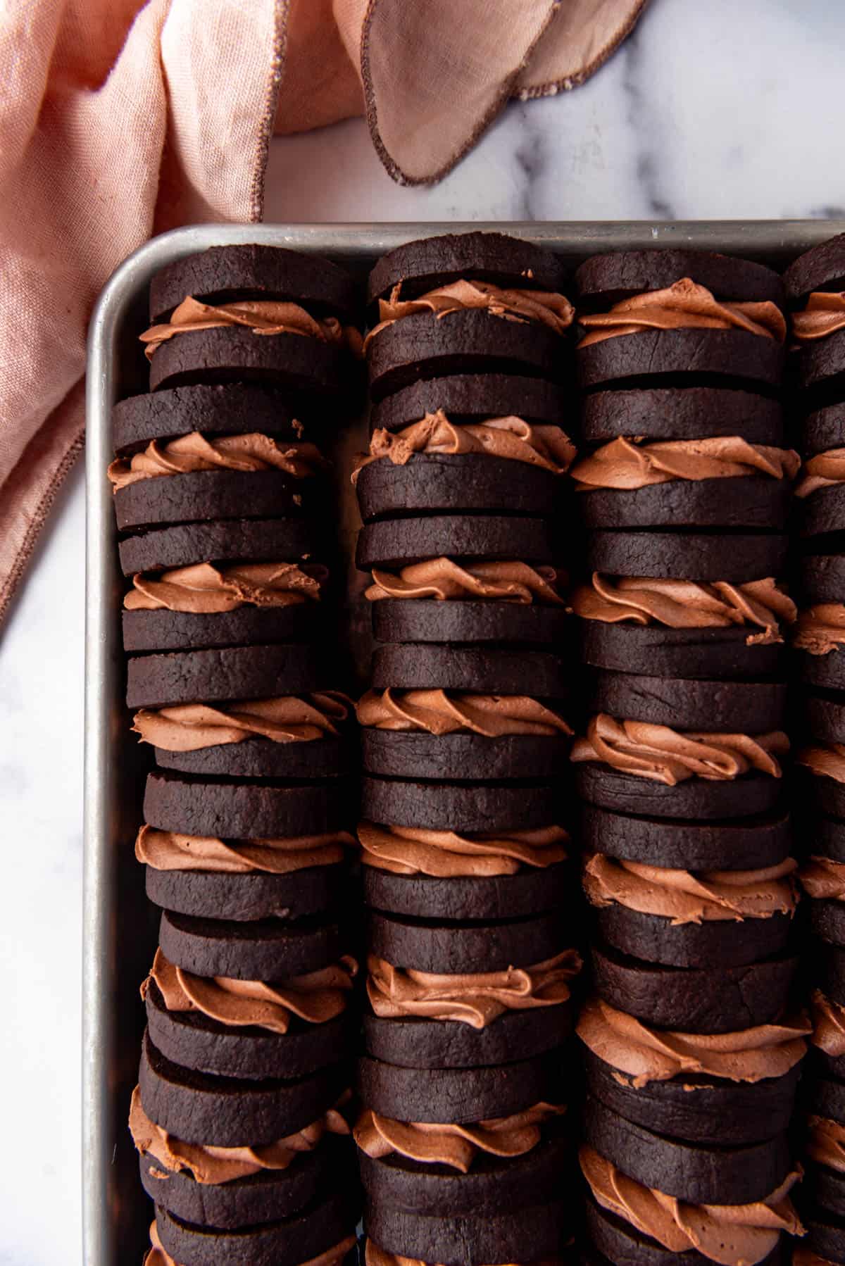 An overhead image of chocolate nutella sandwich cookies stacked in rows on a baking sheet next to a pink cloth napkin.