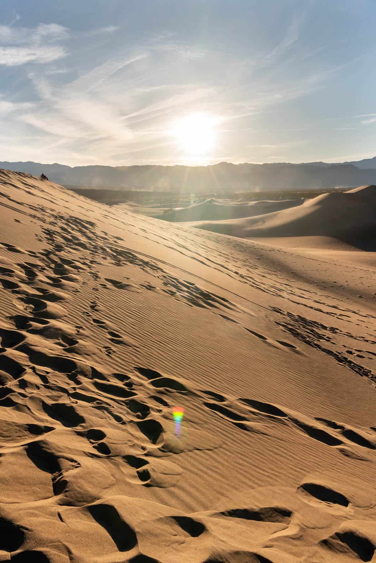 The sun setting over the mountains with a sand dune covered in footprints in the foreground.