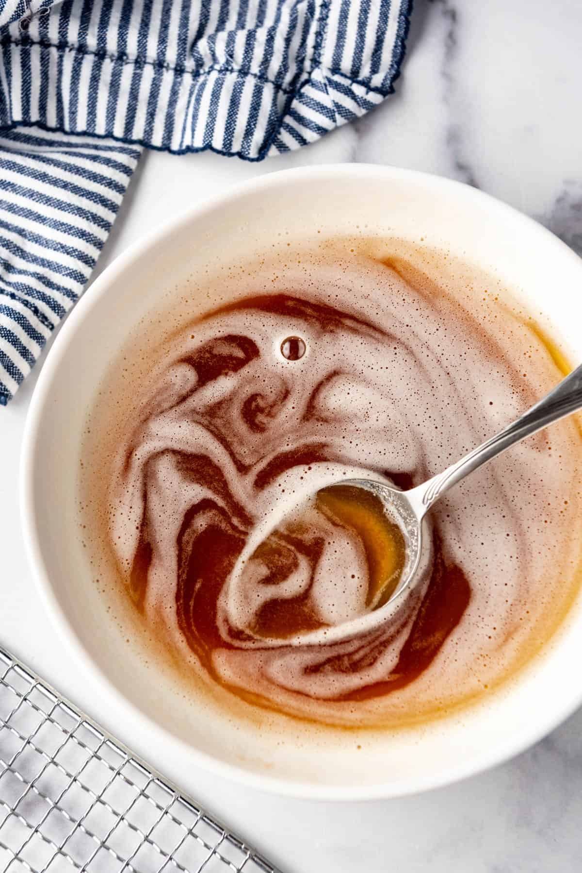 An overhead image of a bowl of brown butter with a spoon in it next to a striped cloth napkin and a wire rack.