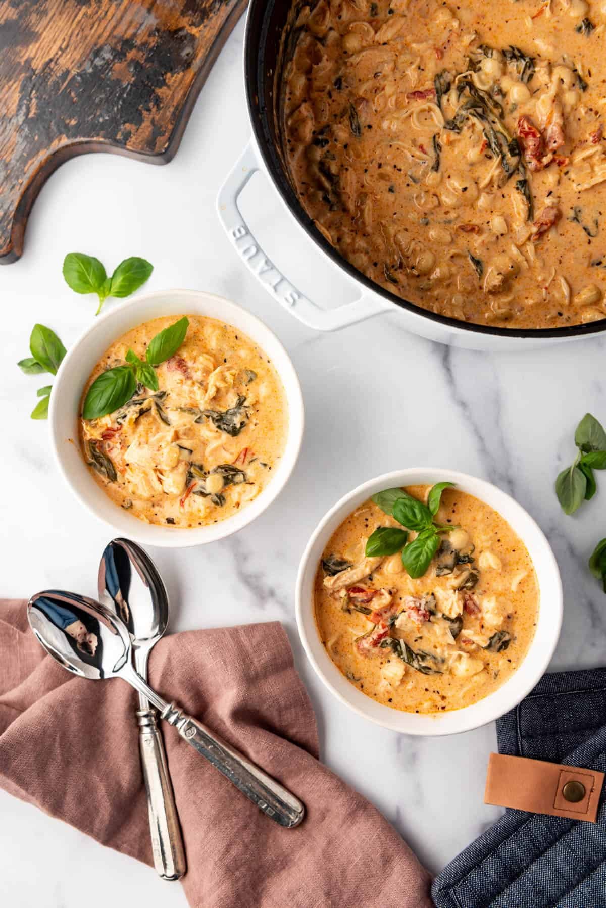 An overhead image of two bowls of marry me chicken soup next to the pot of soup, a cloth napkin, spoons, and a cutting board.