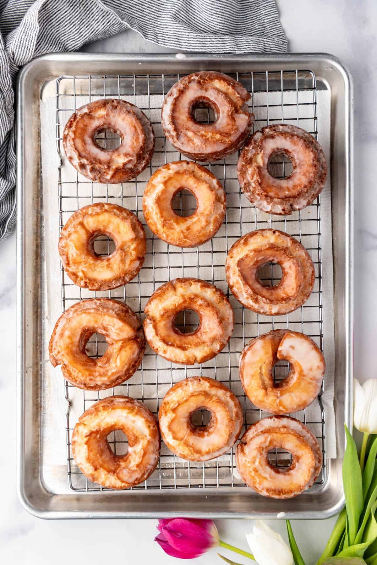 An overhead image of a dozen old-fashioned doughnuts on a wire rack next to some tulips.