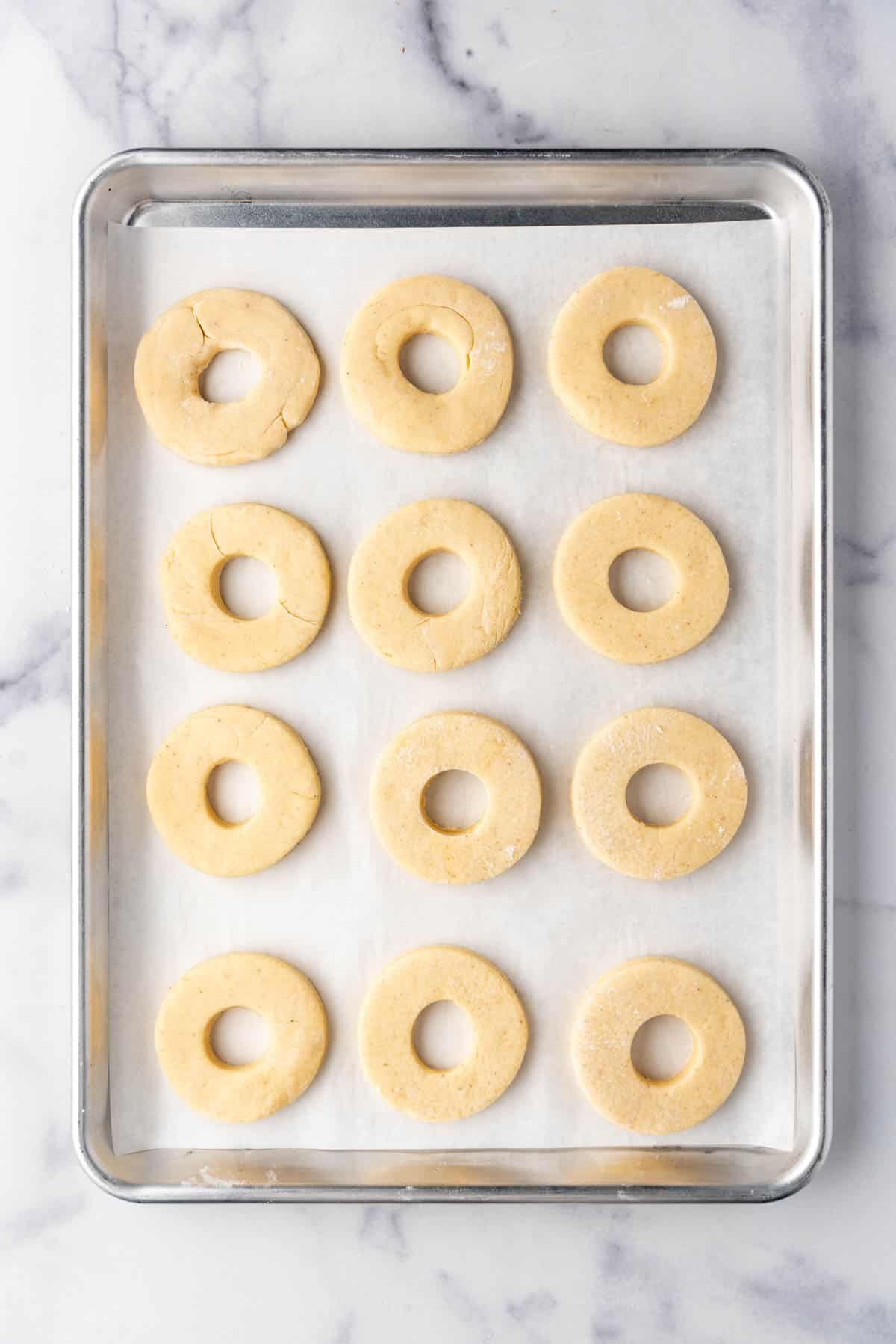 An overhead image of 12 uncooked doughnuts on a baking sheet lined with parchment paper.