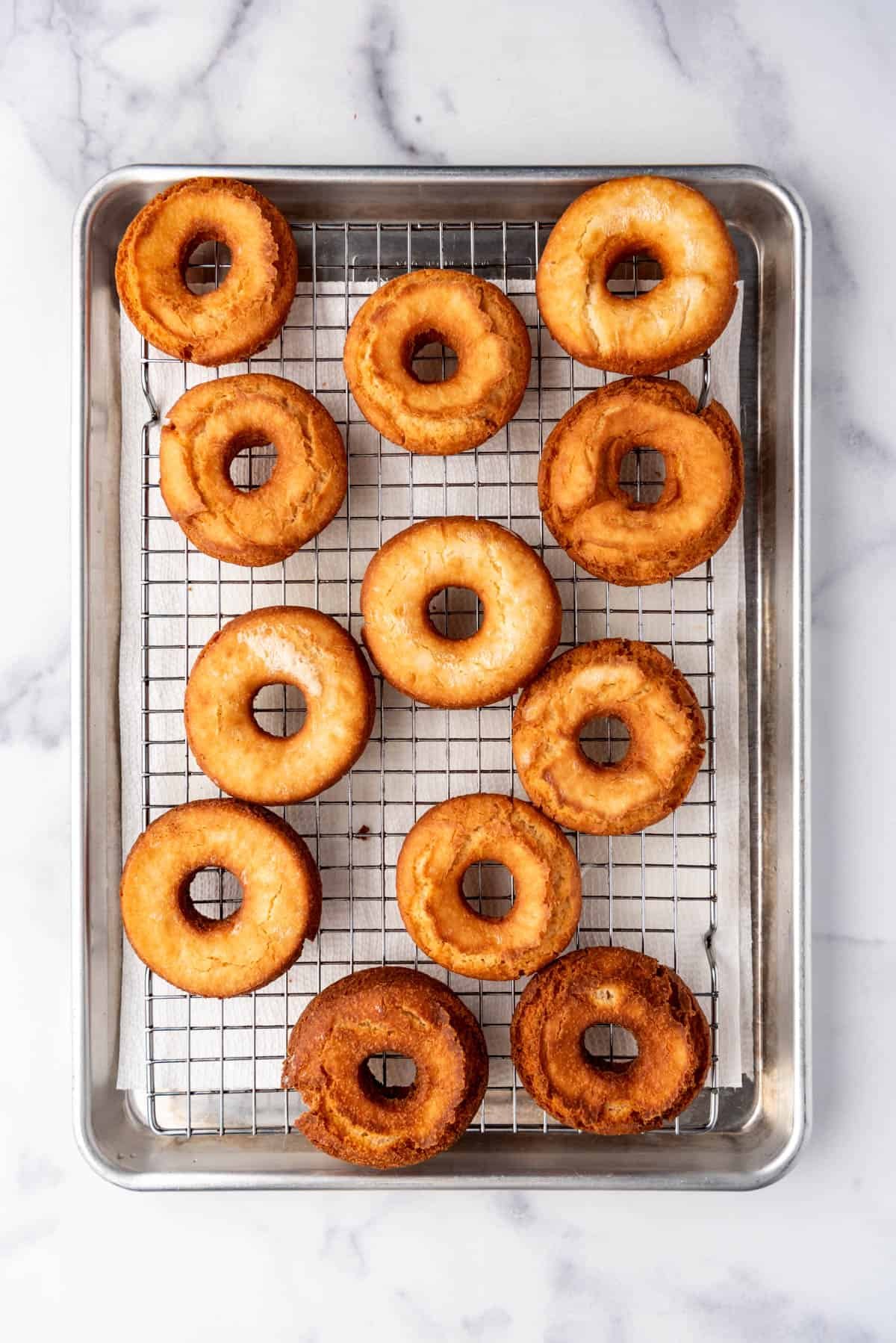 An overhead image of fried doughnuts before glazing on a wire rack set over a baking sheet.