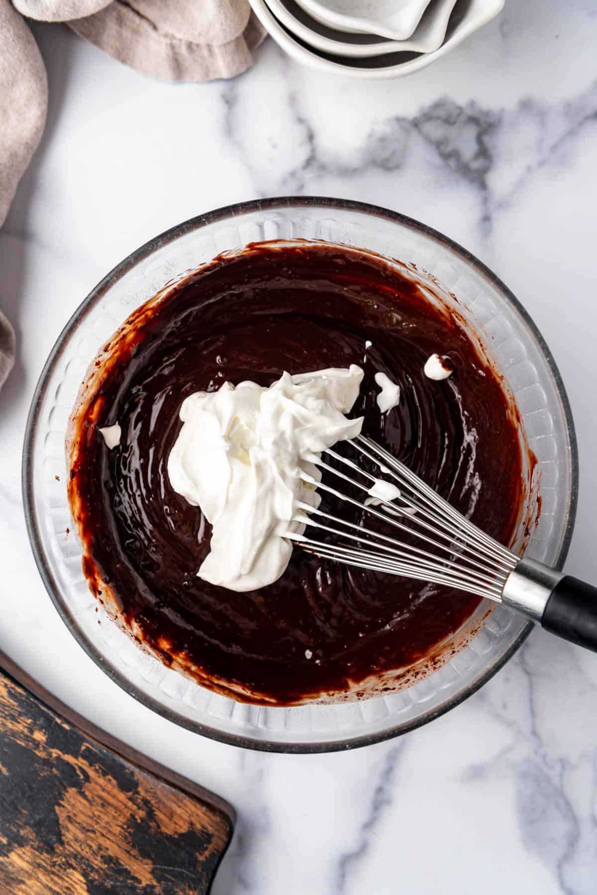 Adding sour cream to fudge frosting in a mixing bowl.