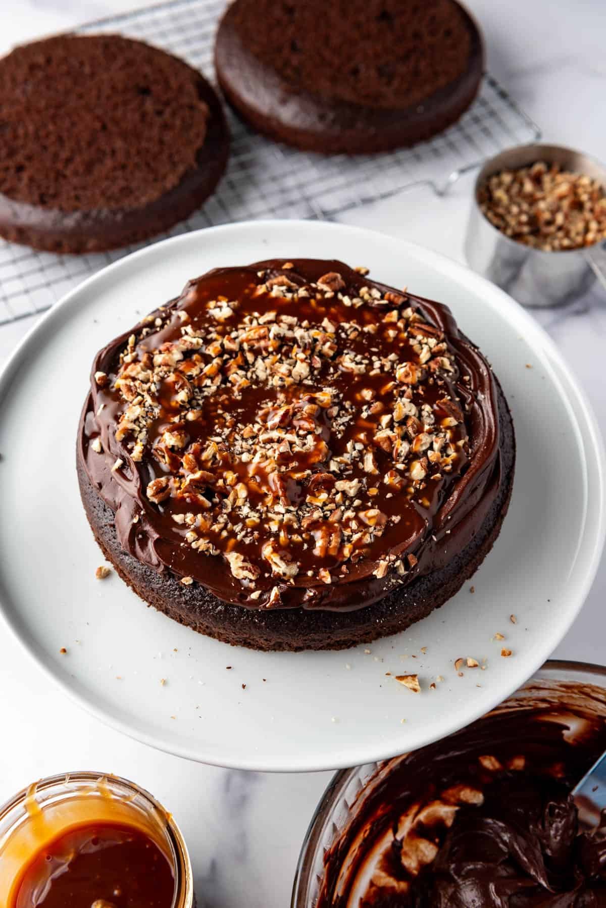 Layering fudge frosting, homemade caramel sauce, and chopped pecans on a layer of chocolate cake on a white cake plate.