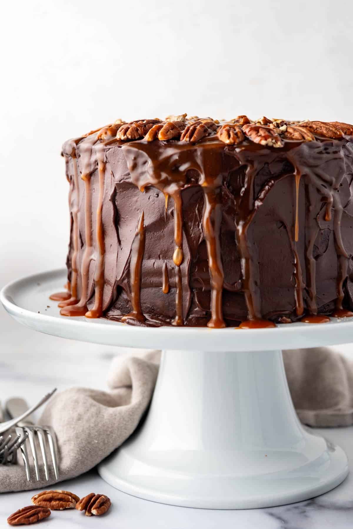 A side angle of a chocolate caramel turtle cake on a white cake stand.