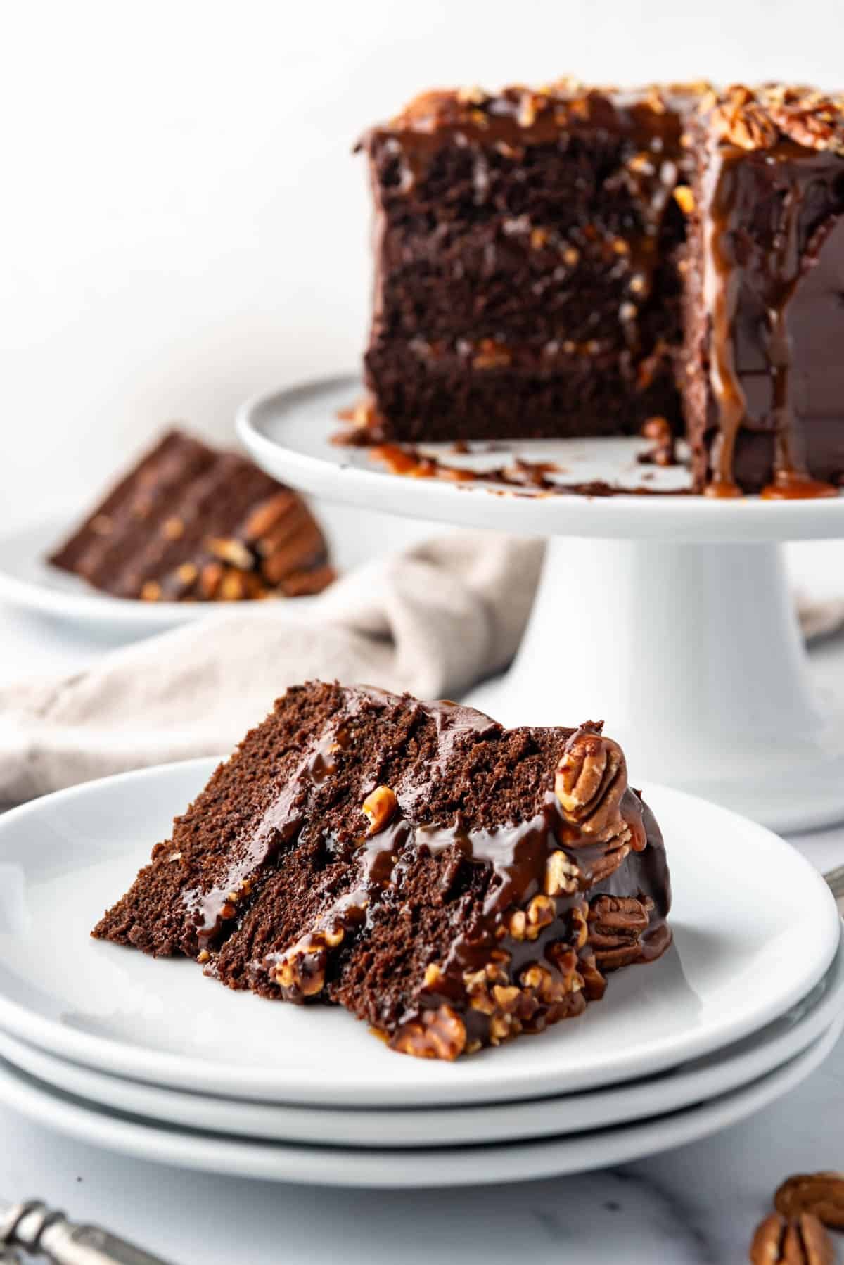 A slice of turtle cake on a stack of three white plates in front of the rest of the cake on a cake stand.