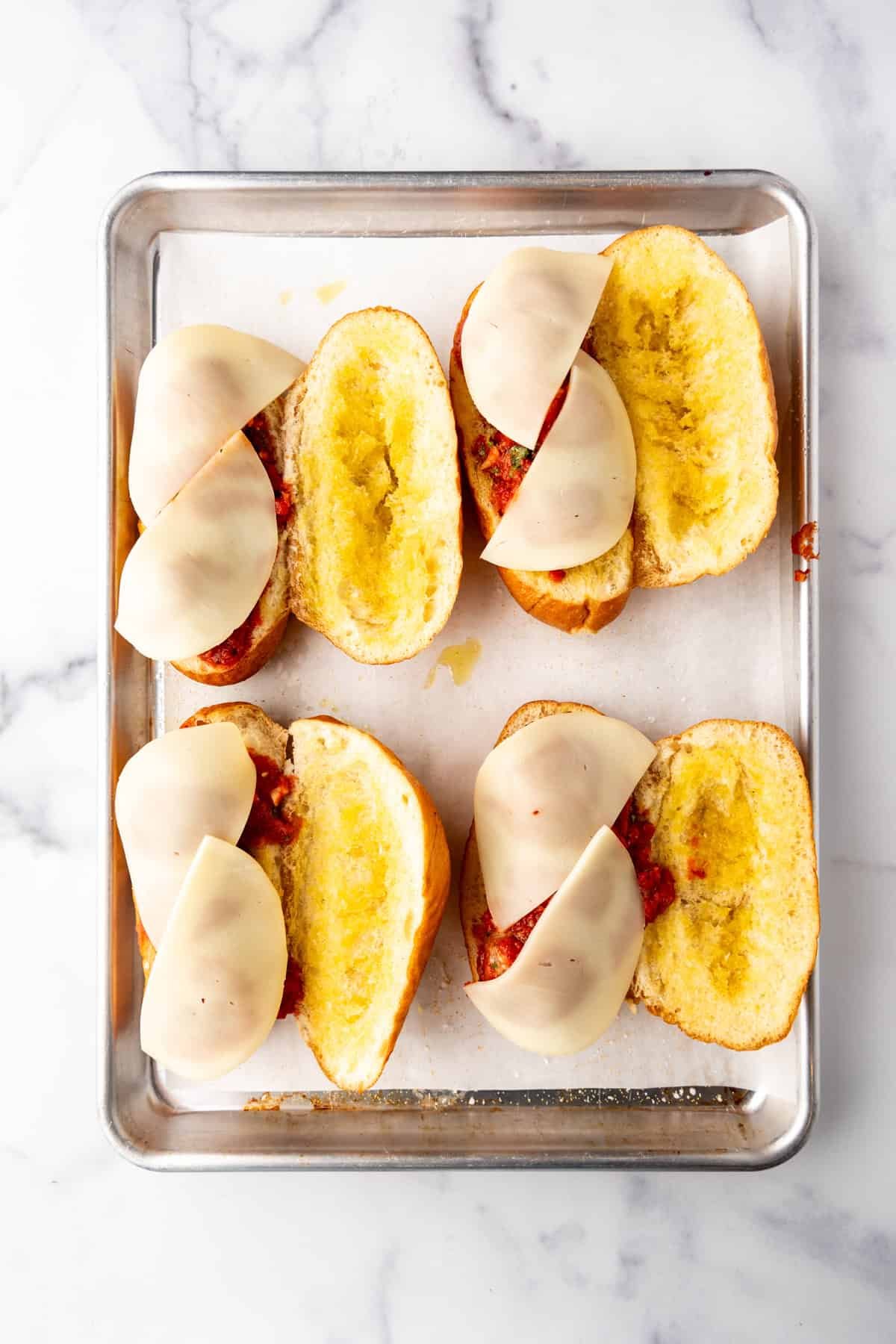 Adding Italian meatballs and sliced provolone cheese to toasted buns on a baking sheet.