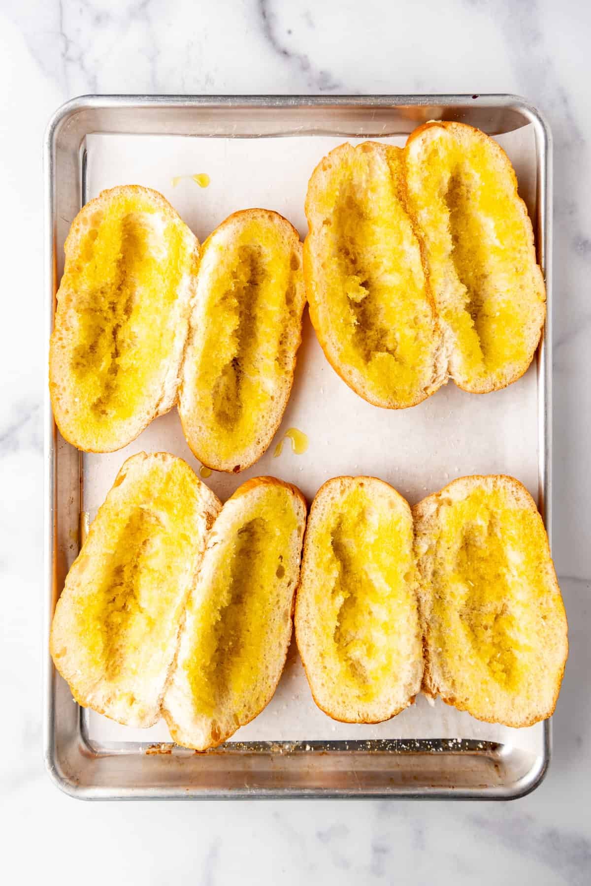 Crusty rolls brushed with olive oil on a baking sheet.