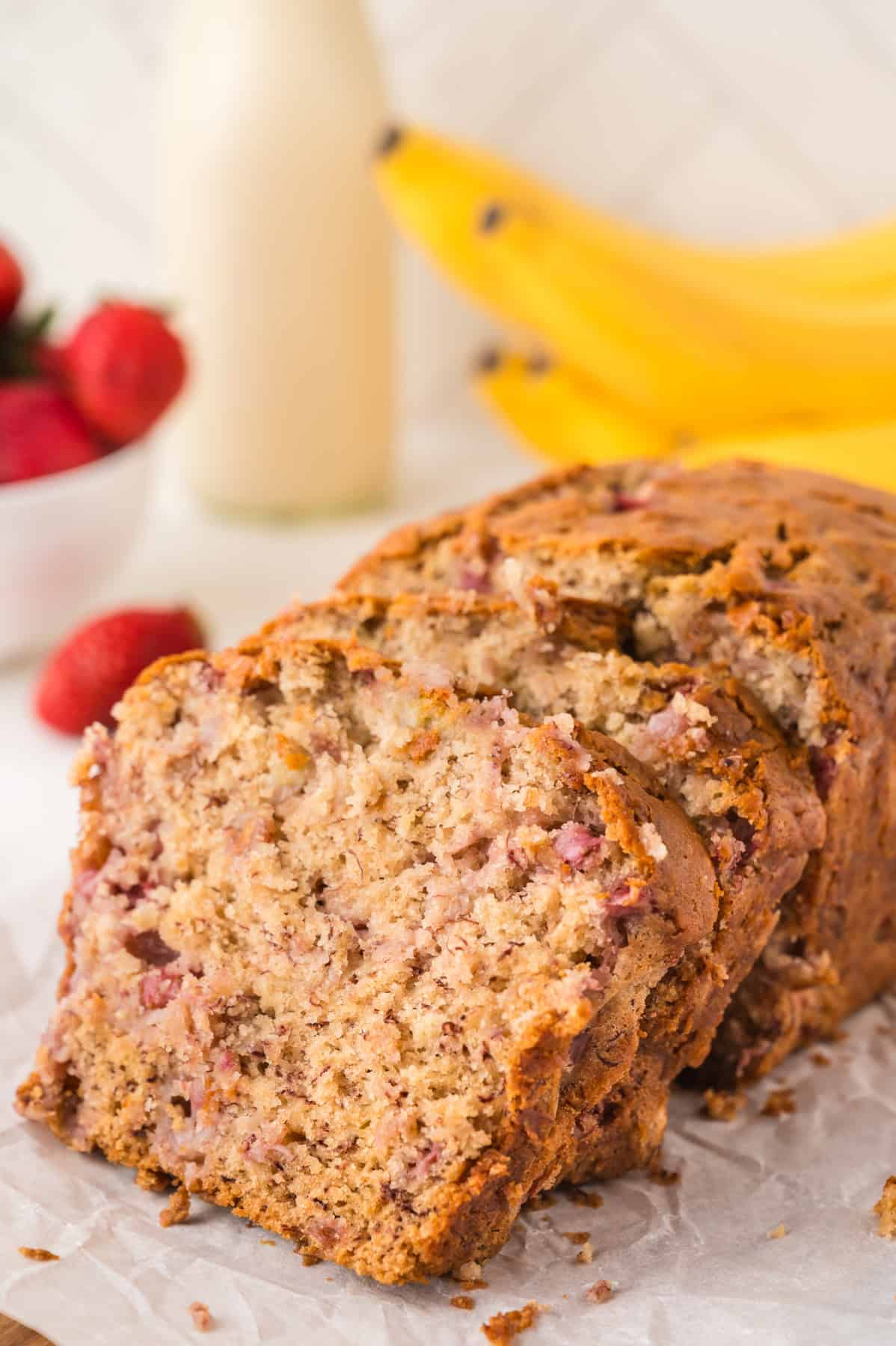 A sliced loaf of strawberry banana bread with bananas and strawberries in the background.