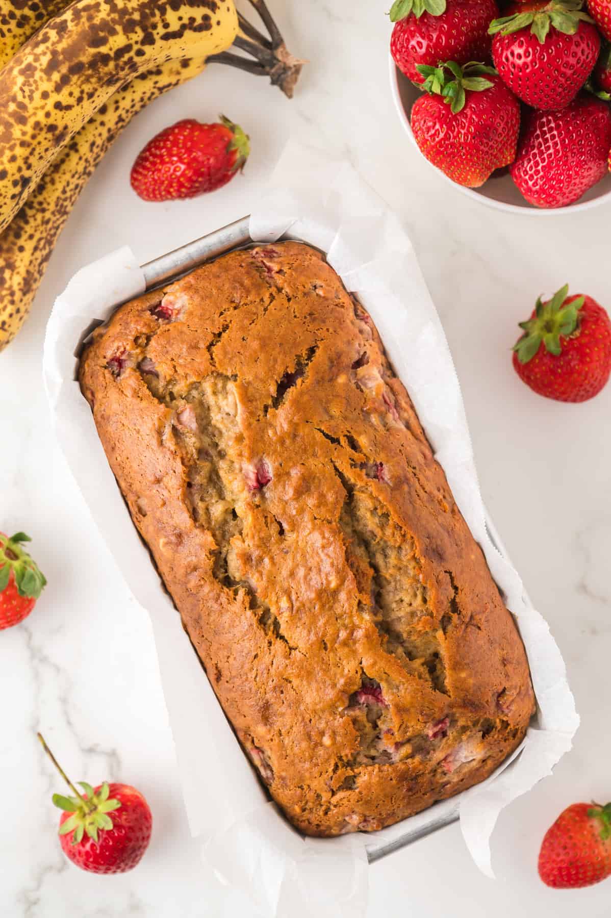 A loaf of strawberry banana bread surrounded by fresh strawberries and spotted bananas.
