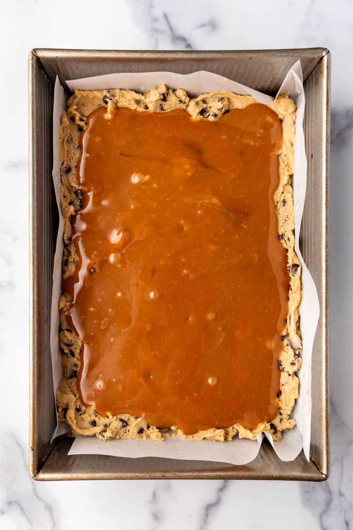 Adding a layer of melted caramel to chocolate chip cookie dough in a 9x13-inch pan.