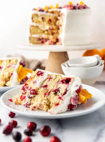 A slice of cranberry orange cake on a white plate with fresh cranberries scattered in front.