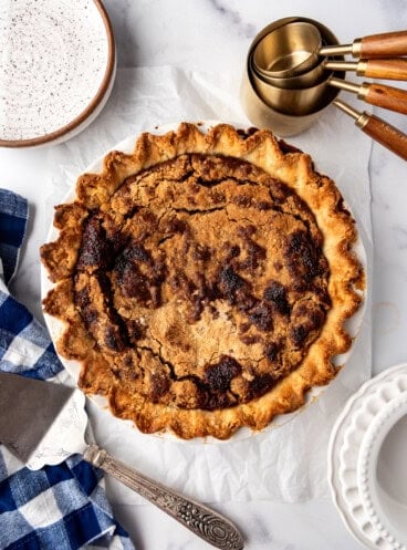 An overhead image of a shoo fly pie surrounded by plates, measuring cups, and a pie server.