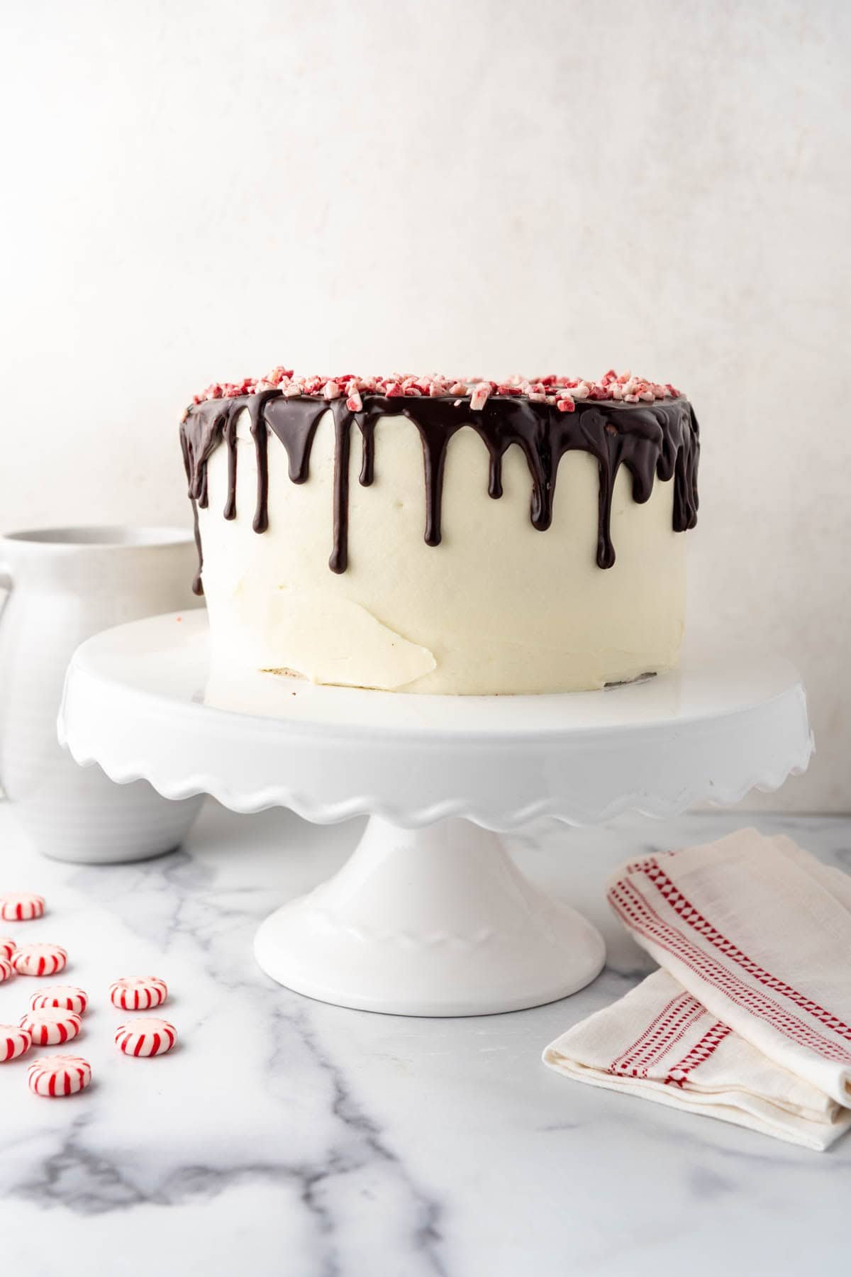 An image of a homemade peppermint bark cake on a white cake stand with peppermint candies and a red and white linen napkin nearby.