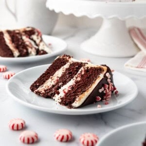 Slices of peppermint bark cake on white plates in front of the rest of the cake on a cake stand.