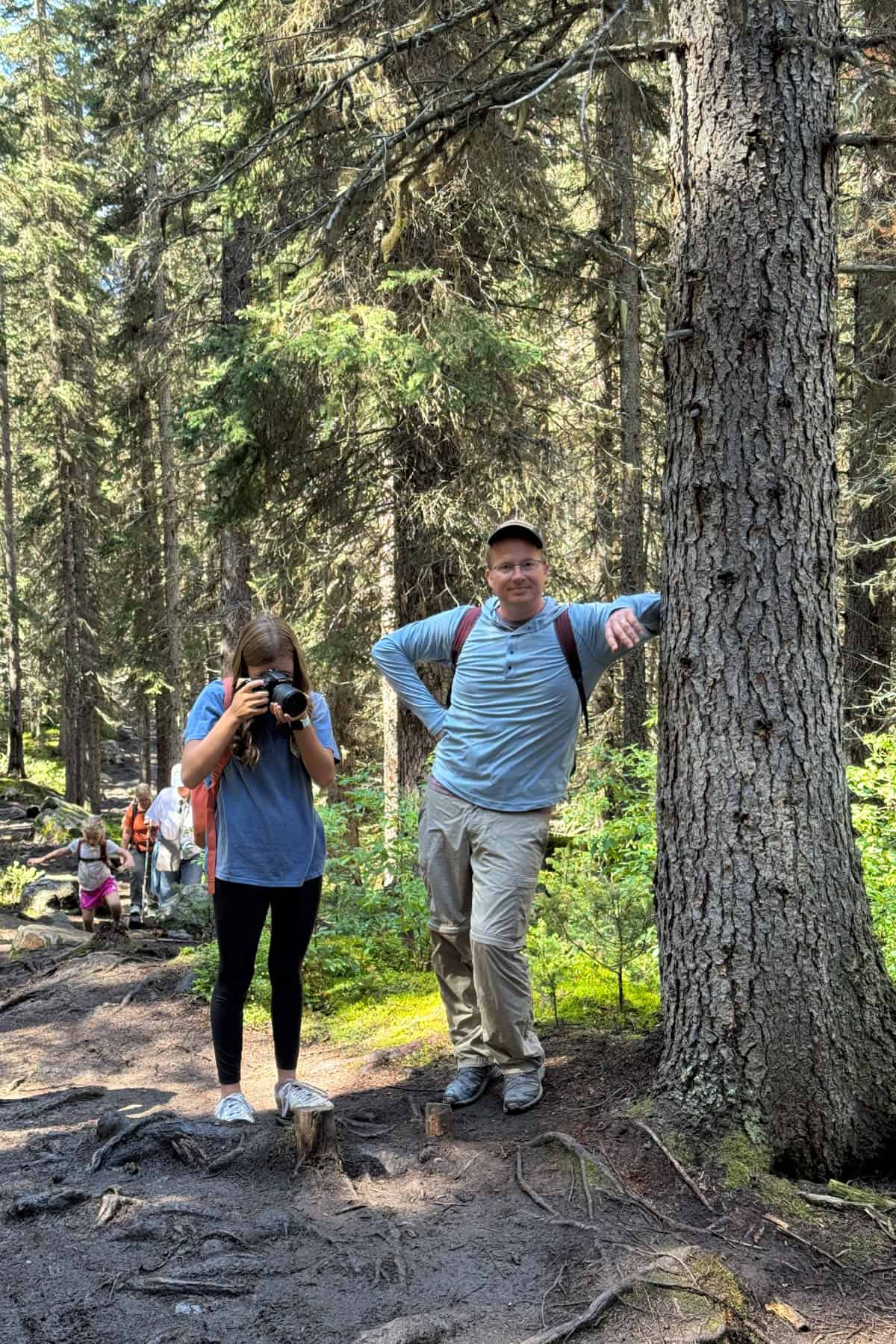 A man leaning against a tree while a girl takes pictures of the tree roots.