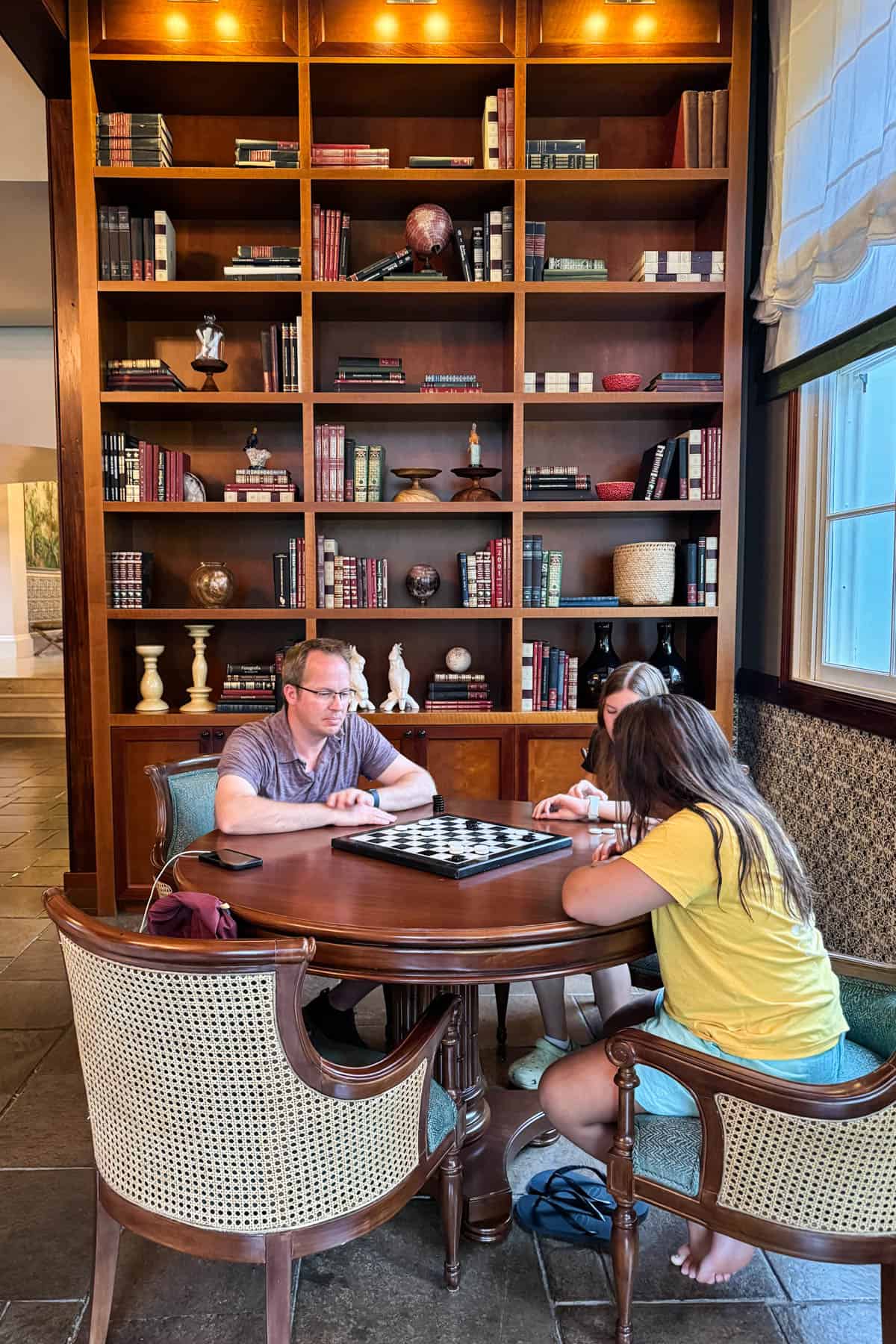 A dad and kids playing checkers at a game table with a bookshelf behind them.