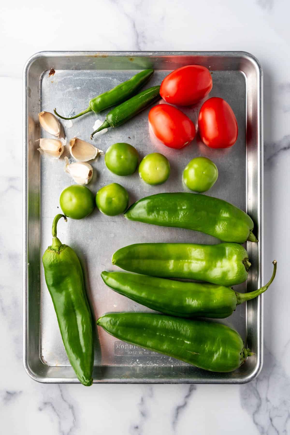 Fresh peppers, tomatillos, tomatoes, and garlic cloves on a baking sheet.