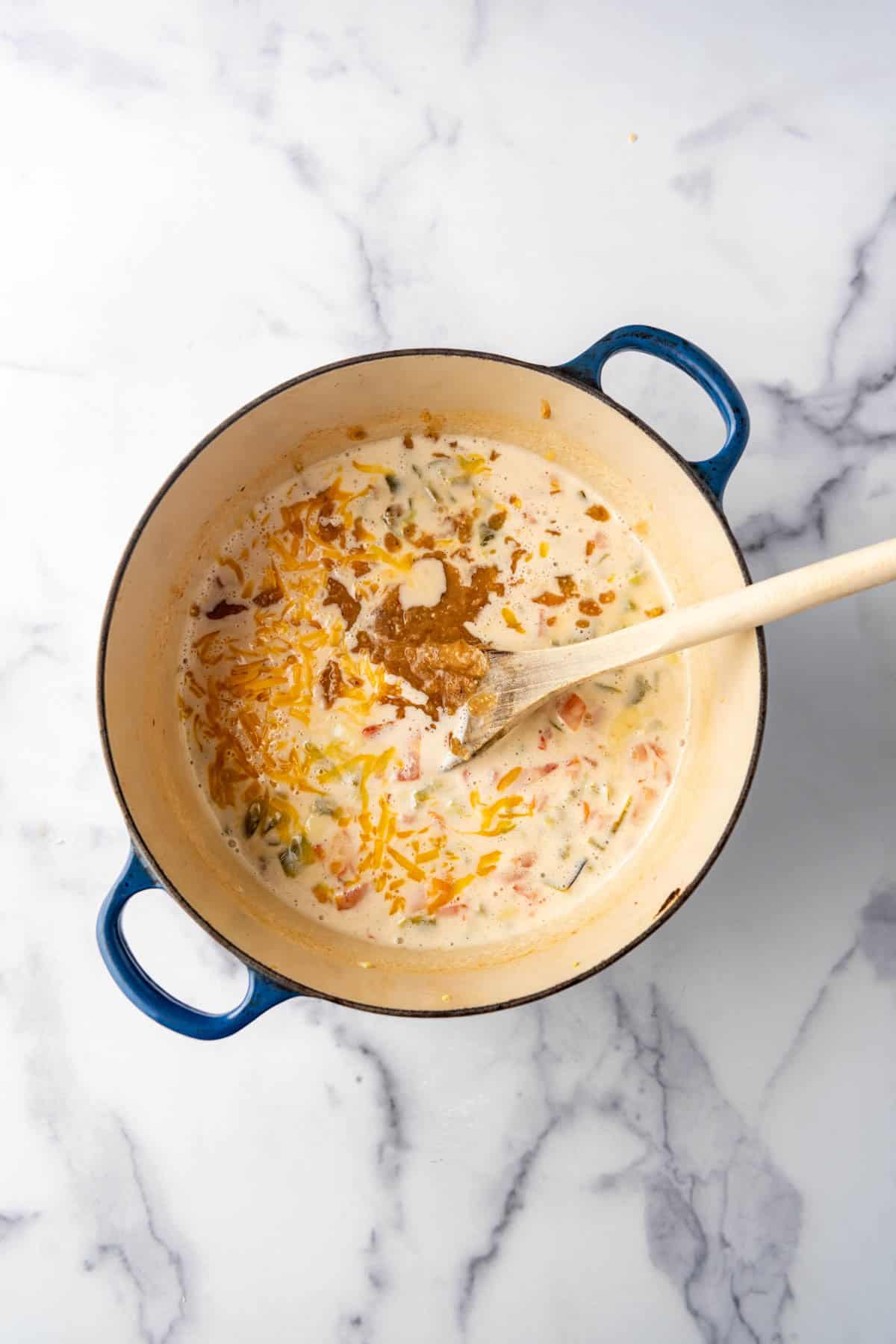 An overhead image of a pot of queso with grated cheddar cheese and salsa verde being stirred in with a wooden spoon.