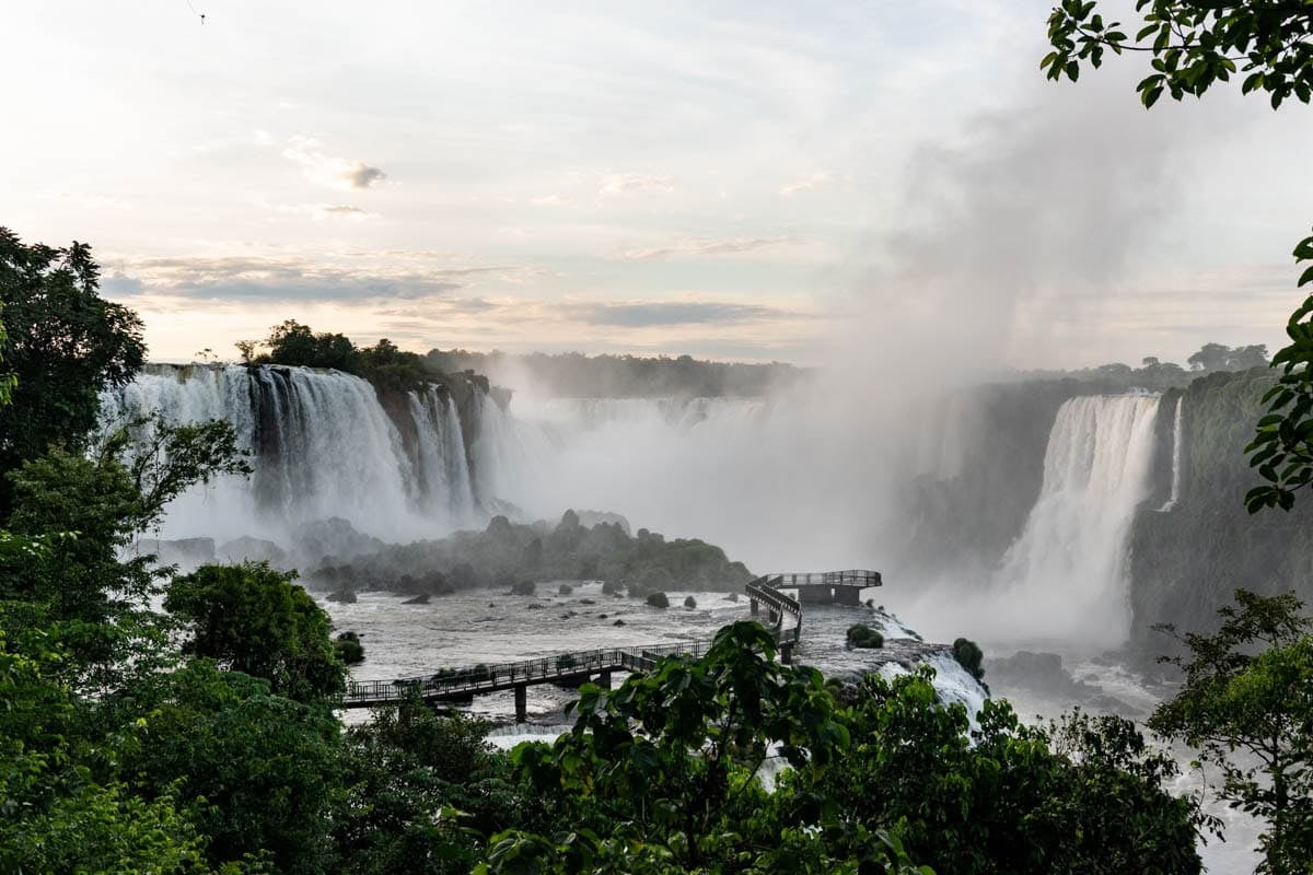 A dawn view of mist rising up from Iguazu Falls in Brazil with the walkway and viewing platform in the foreground.