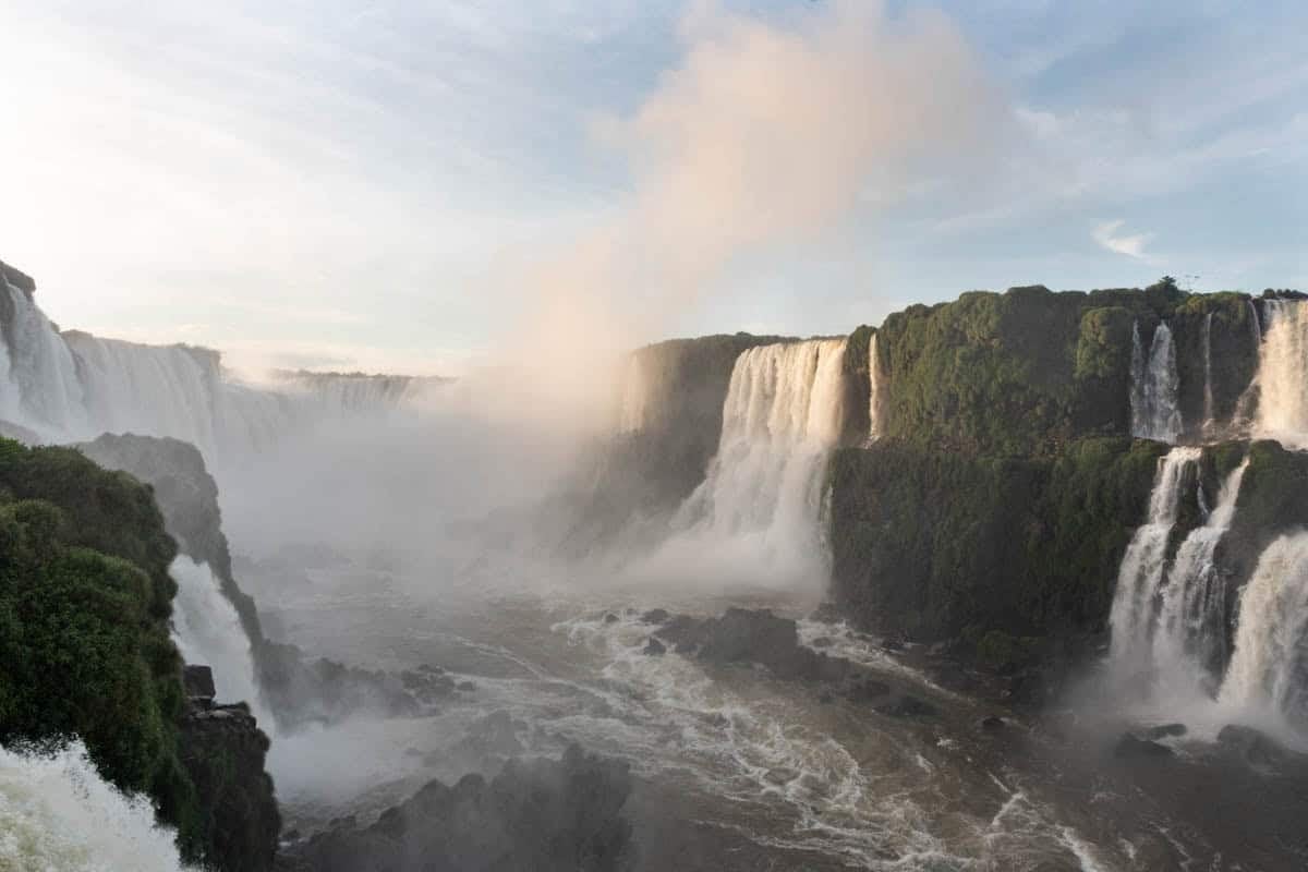 A panoramic view of mist rising from Iguazu Falls in early morning light.